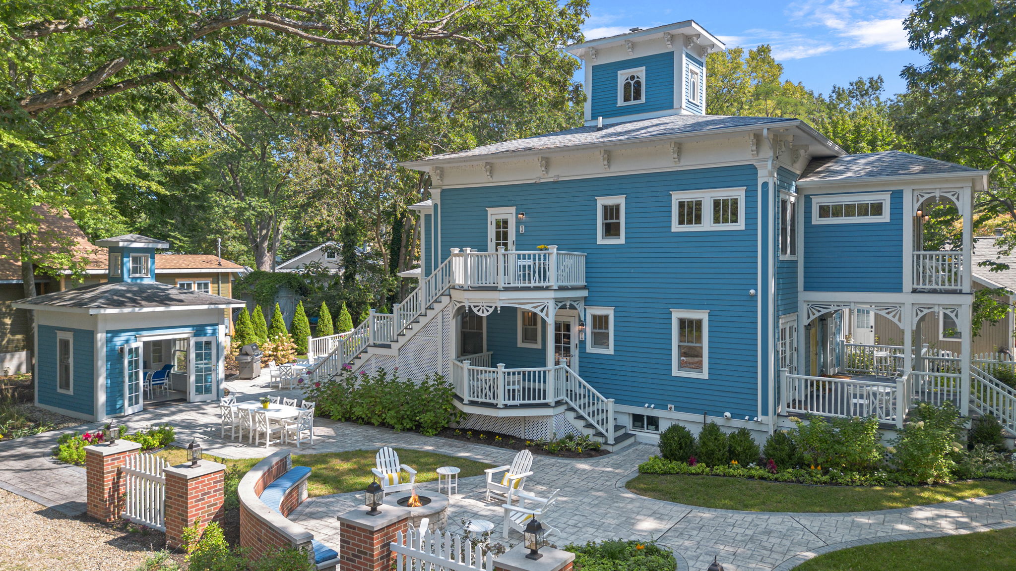 An elevated, wide-angle view of the Union Pier Summer House, a large three-story home with vibrant blue siding and ornate white trim. The property features extensive white-railed porches and a beautifully landscaped yard with stone pathways, a circular fire pit area with white Adirondack chairs, and a matching blue garden pavilion.