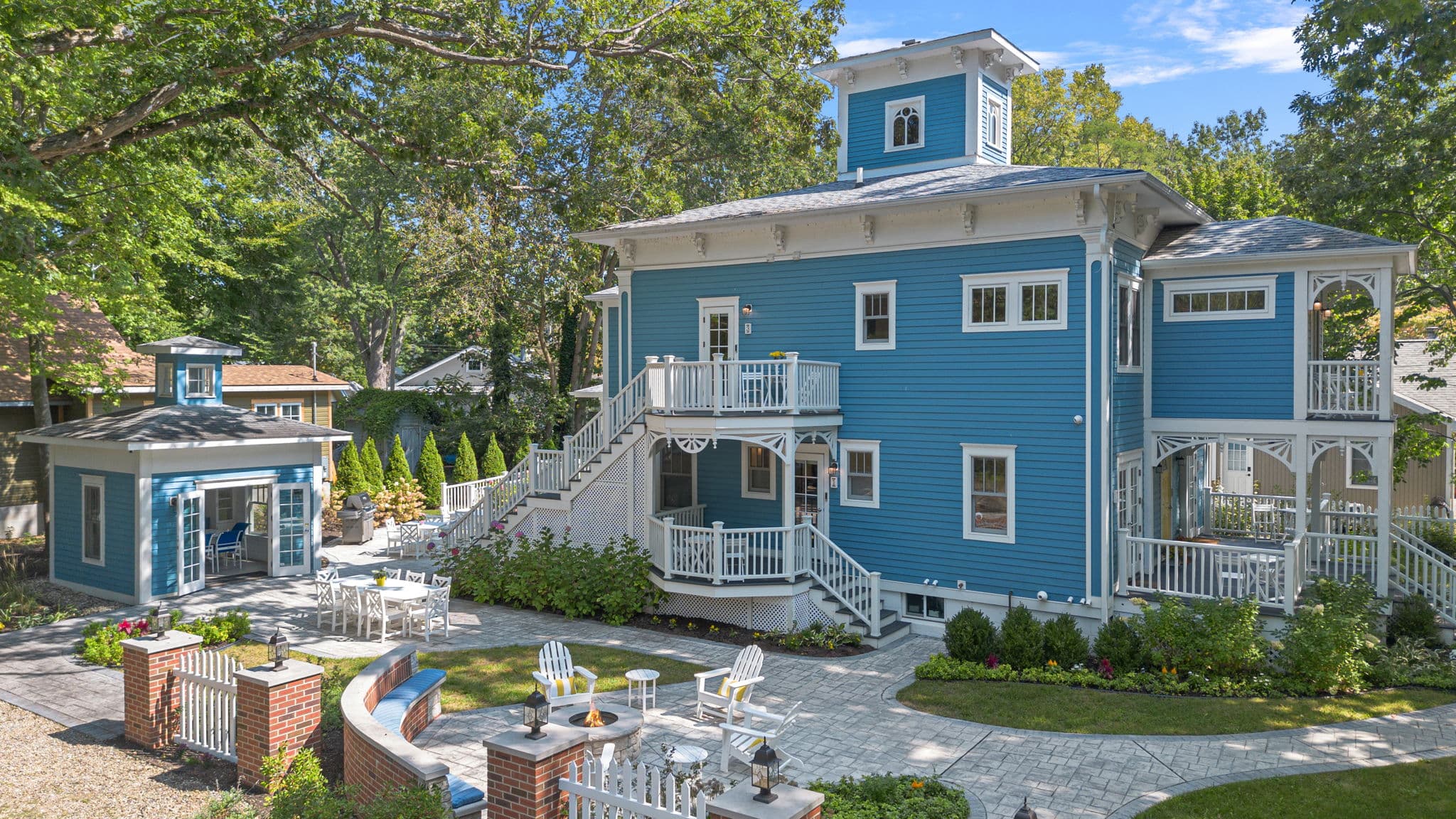 An elevated, wide-angle view of the Union Pier Summer House, a large three-story home with vibrant blue siding and ornate white trim. The property features extensive white-railed porches and a beautifully landscaped yard with stone pathways, a circular fire pit area with white Adirondack chairs, and a matching blue garden pavilion.