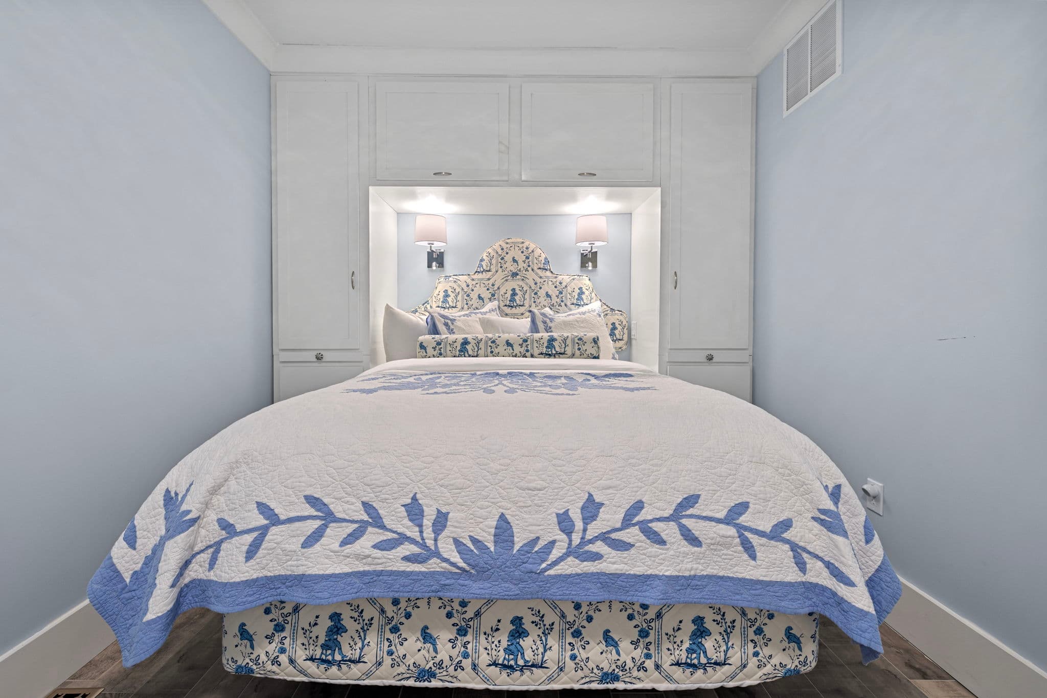 A bright, narrow bedroom with light blue walls featuring a queen bed tucked into a custom white storage unit with built-in closets and overhead cabinets. The bed is dressed in a white quilt with a large blue floral pattern, matching a decorative headboard.