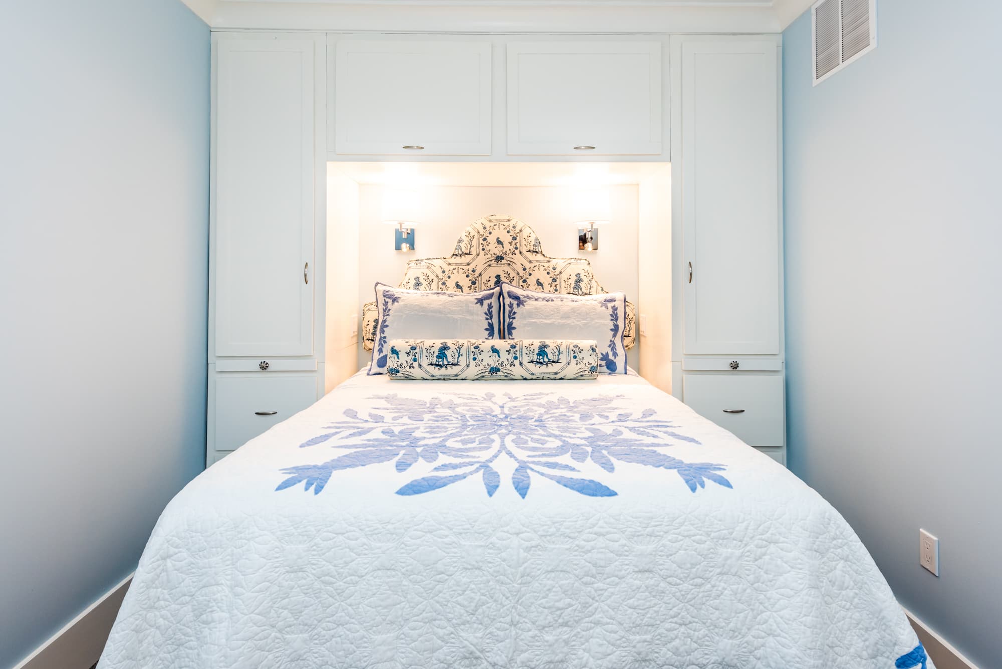A bright, narrow bedroom with light blue walls featuring a queen bed tucked into a custom white storage unit with built-in closets and overhead cabinets. The bed is dressed in a white quilt with a large blue floral pattern, matching a decorative headboard.