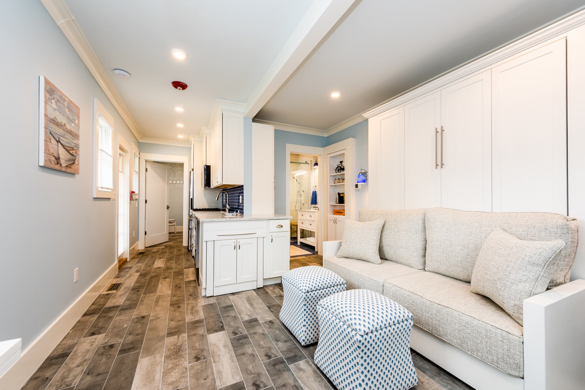 A bright, modern living area with light blue walls and wood-look flooring, featuring a wall-mounted TV above a white desk and chair, and glass doors that open onto a sunlit outdoor deck.