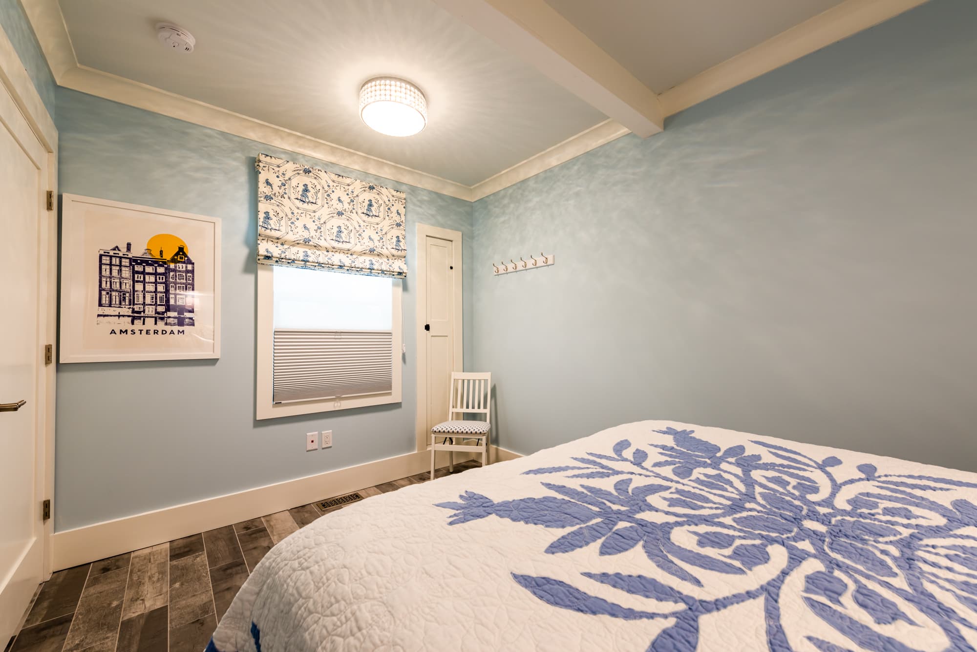 A bright bedroom with light blue walls and wood-look flooring, featuring a bed with a blue and white floral quilt, a matching patterned window shade, and a framed art print of a cityscape.