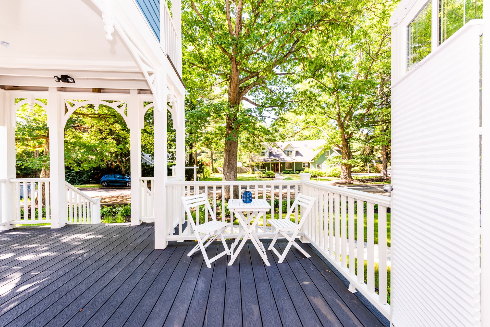 A bright, elevated outdoor south-facing front deck with dark grey floorboards and white railings, featuring a small white bistro table and two matching folding chairs overlooking a lush green yard with mature trees.