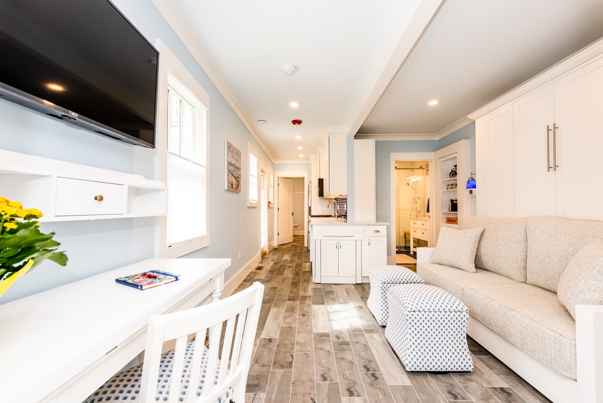 An airy, modern living area with light blue walls and wood-look flooring, featuring a white Murphy sofa bed with light blue ottomans, a built-in desk with a flat-screen TV above it, and a view into a bright white kitchen.
