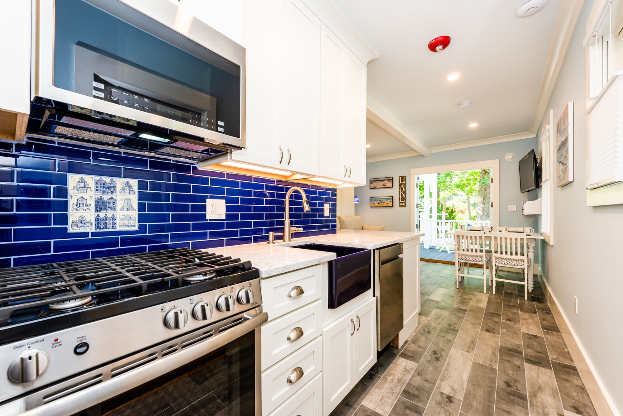A modern kitchen with white cabinetry and a vibrant blue subway tile backsplash, featuring a stainless steel gas range, a farmhouse sink with a gold faucet, and a view into a bright dining area with glass doors leading outside.