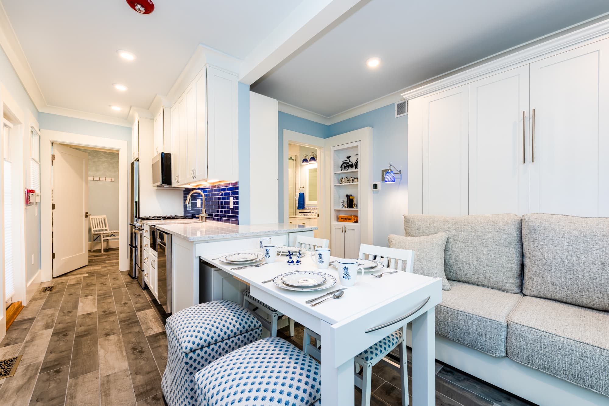 A bright dining and living area featuring a white table set for two with blue-and-white patterned ottomans, adjacent to a white Murphy sofa bed and a modern kitchen with a blue tiled backsplash.