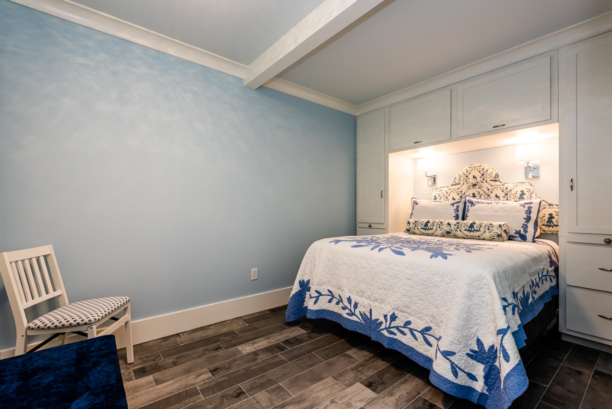 A bright bedroom with light blue walls and wood-look flooring, featuring a bed tucked into custom white cabinetry and dressed in a white quilt with a blue floral pattern.