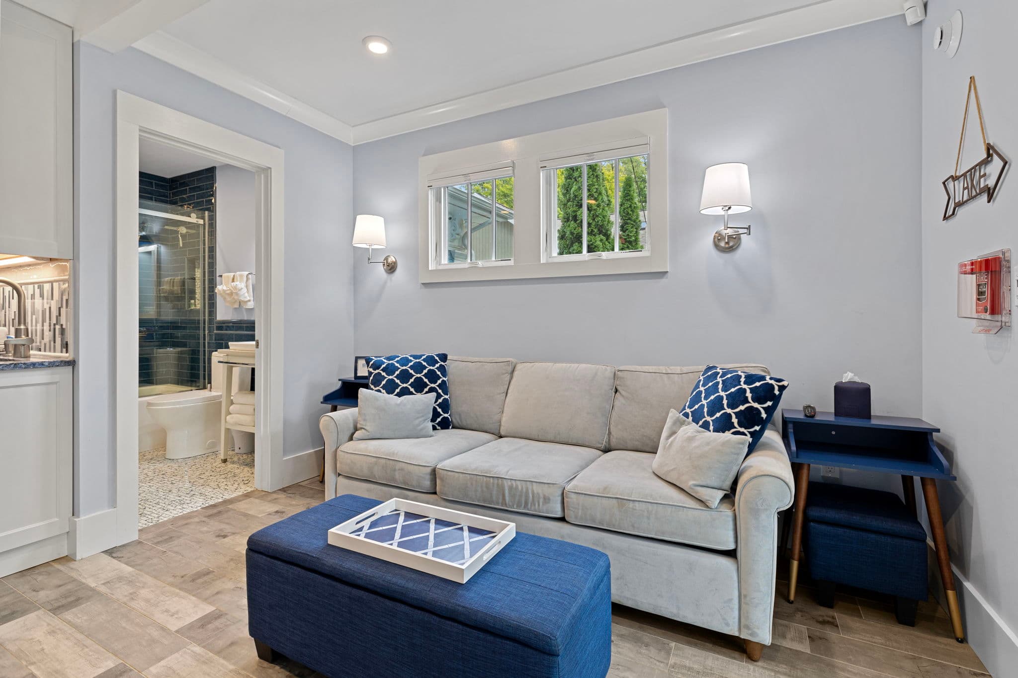 A cozy living area with light blue walls and wood-look tile flooring, featuring a light grey sofa with blue patterned accent pillows and a large navy blue ottoman. To the left, an open doorway reveals a bright bathroom.