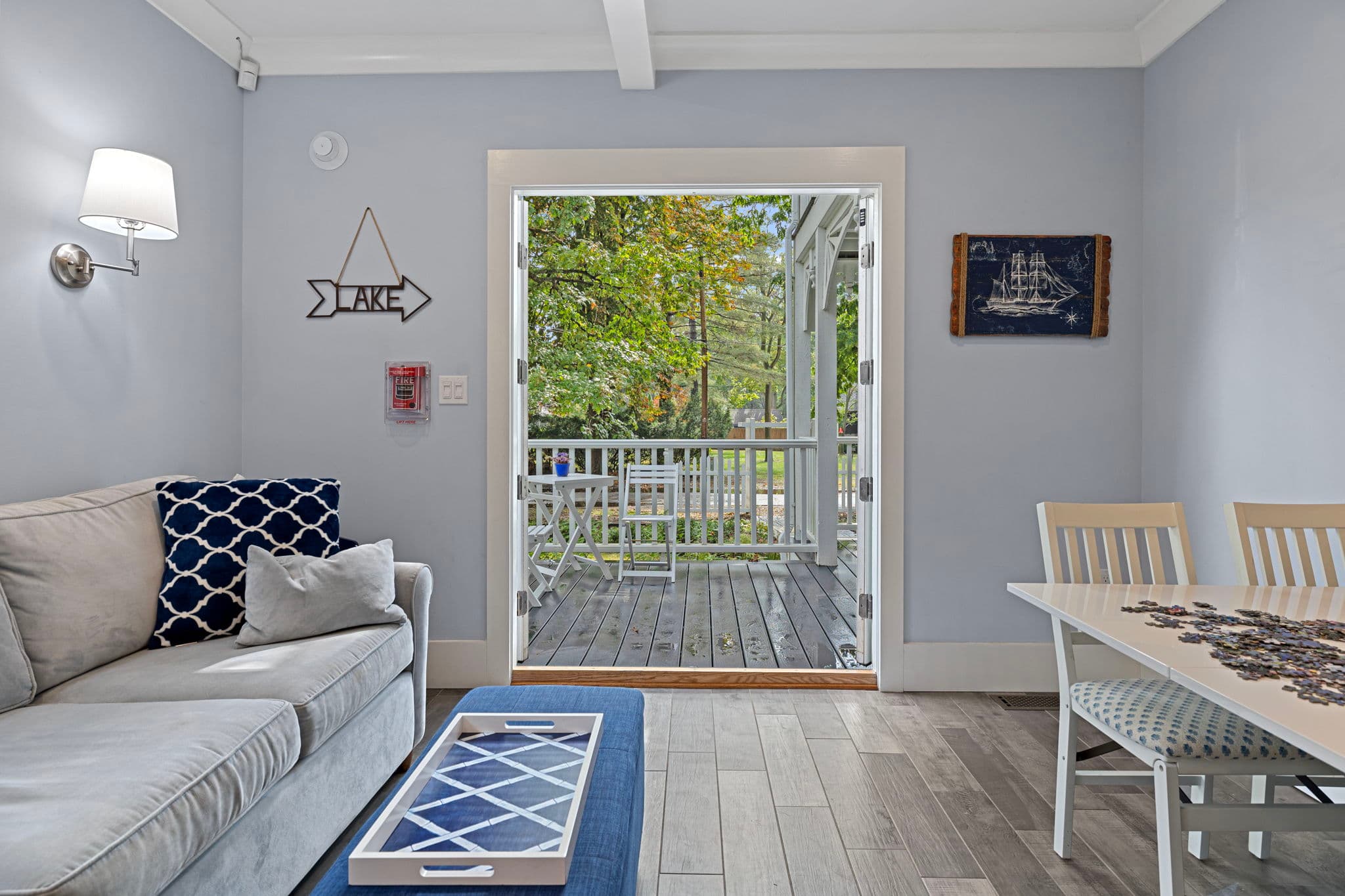 A bright living area featuring a light grey sofa with decorative blue and white pillows, situated next to a doorway that opens onto a wooden deck overlooking lush green trees. To the right, a portion of a white dining table is visible, all set against light blue walls and wood-plank flooring.