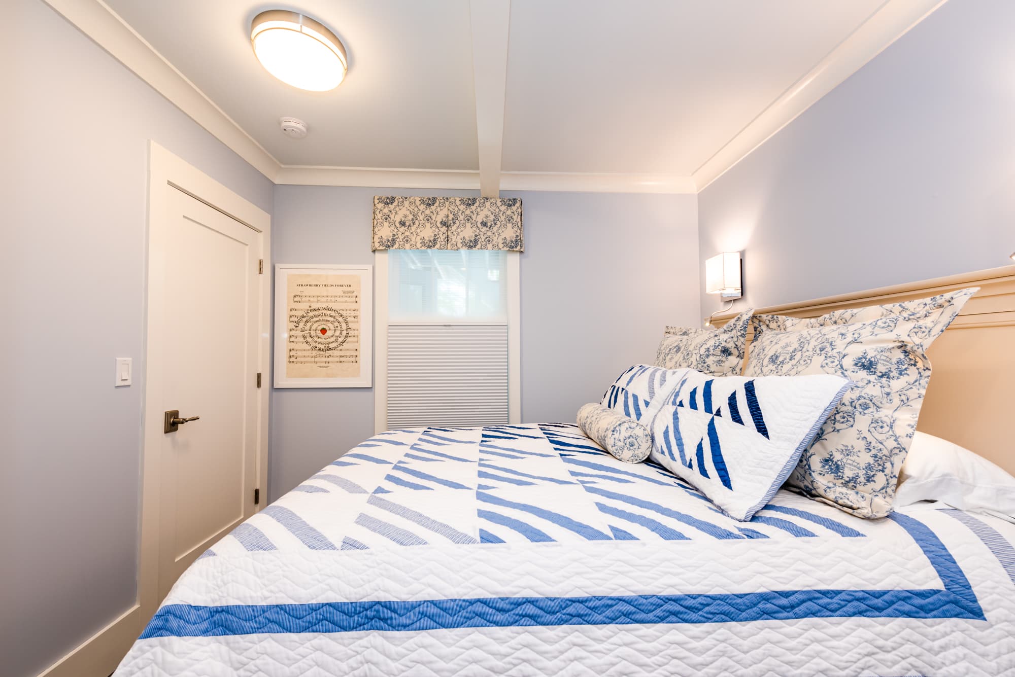 A close-up view of a bed with a vibrant blue and white patterned quilt and matching pillows, set against light blue walls in a bright room with a classic white door and a framed art print on the wall.