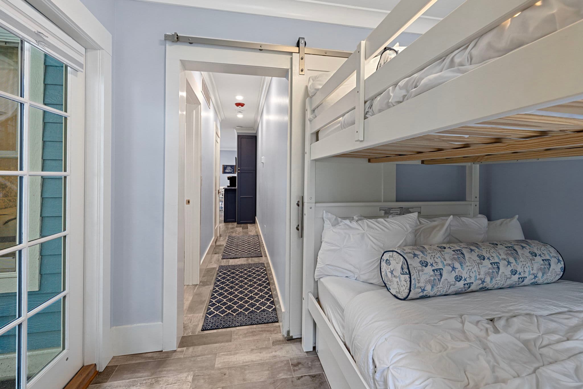 A cozy bedroom with light blue walls featuring white wooden bunk beds tucked into a corner. The lower bunk is dressed in white linens with a blue floral bolster pillow, and a sliding barn-style door leads to a narrow hallway with patterned runners.
