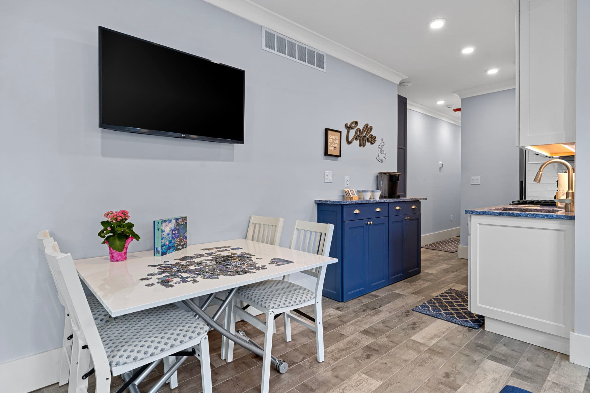 A bright, modern living area with light blue walls and wood-look flooring, featuring a white dining table with two chairs, a wall-mounted LG smart TV, and a blue sideboard with a coffee maker against the far wall.