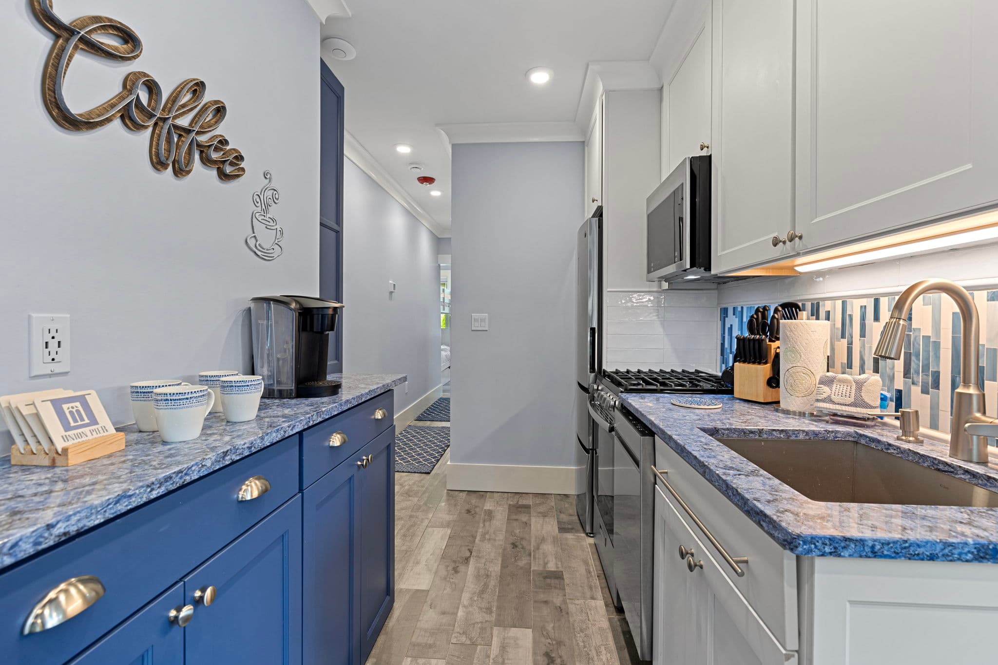 A narrow kitchen space featuring blue and white cabinetry, a blue countertop, and a coffee station area. A stainless steel sink and gas range are visible on the right, while a blue sideboard with a coffee maker and "Coffee" wall decor sits on the left.