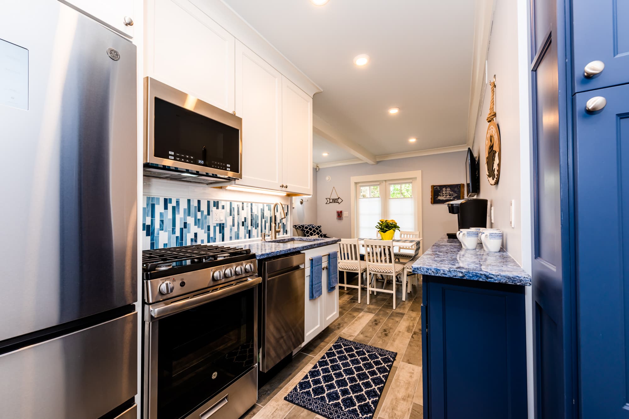 A narrow kitchen featuring white upper cabinets and a stainless steel gas range and microwave. The design includes a striking blue-and-white marble-patterned backsplash and matching countertops, with a large blue storage cabinet and coffee station on the right.