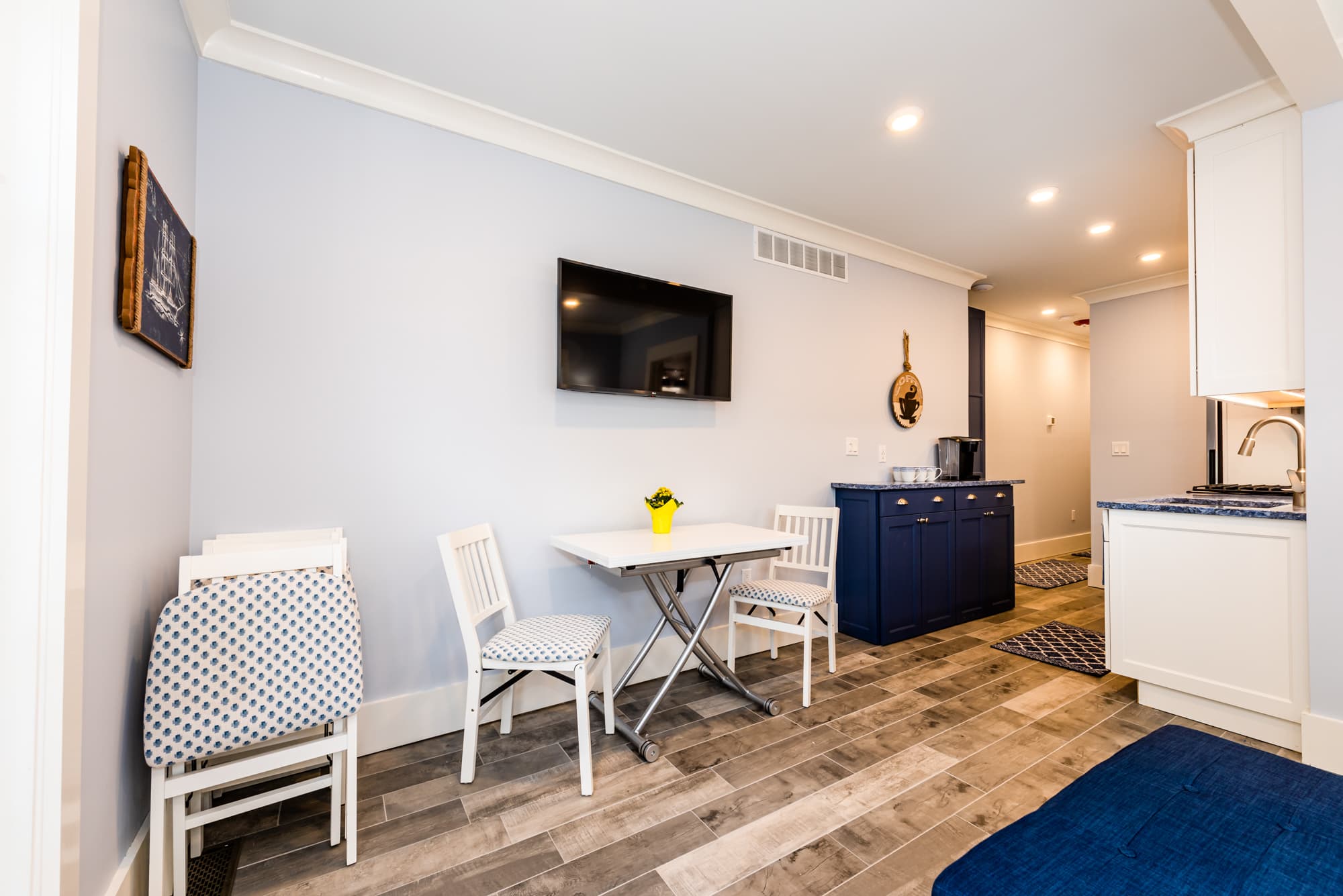 A small, modern dining area and kitchen hallway with light blue walls, wood-plank flooring, and white trim. It features a small white folding table with two white chairs, a wall-mounted TV, and a navy blue sideboard against the far wall.