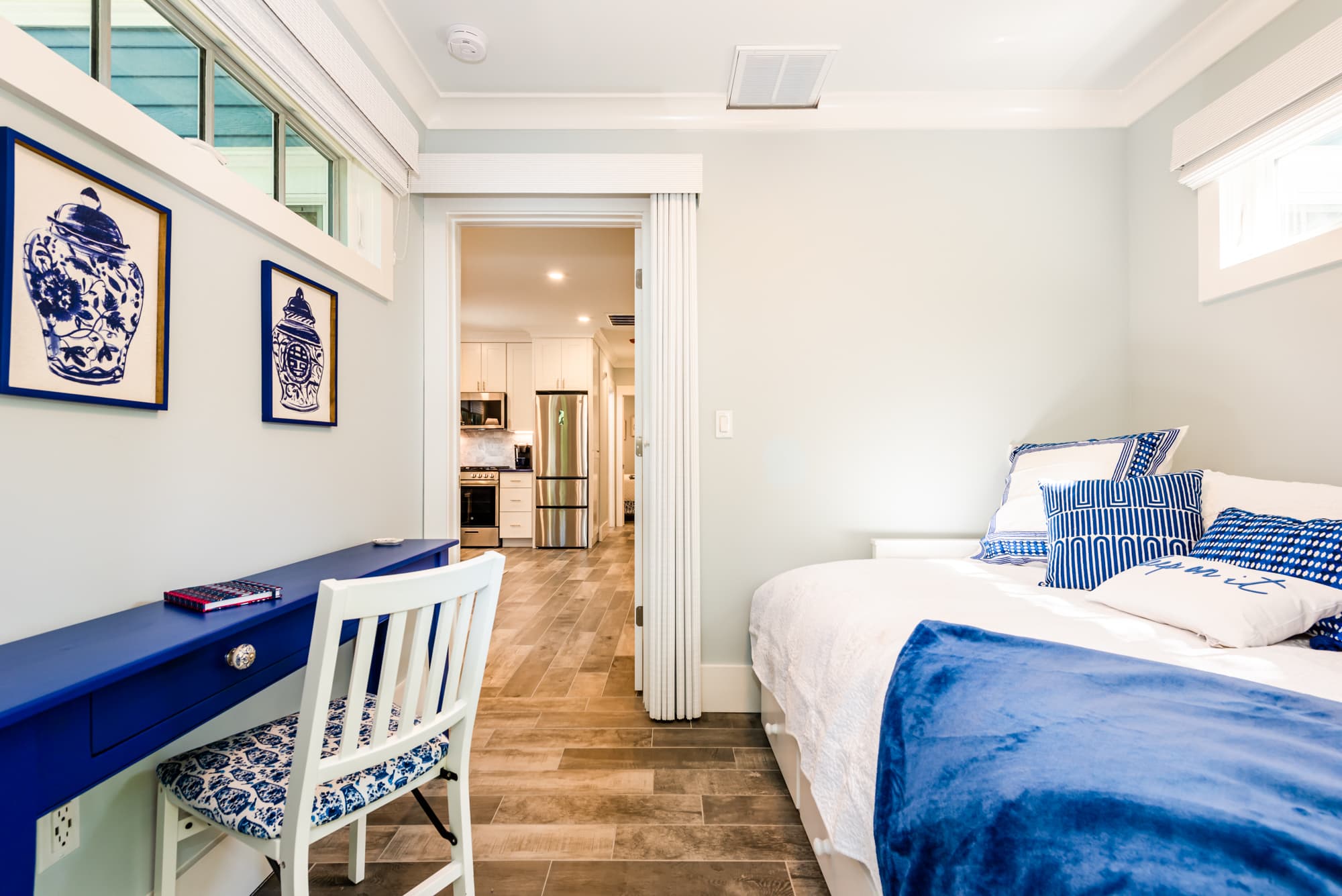 A front bedroom featuring a white bed with blue and white bedding, including a prominent solid blue throw blanket. Against the light blue wall sits a vibrant cobalt blue desk with a matching chair featuring a patterned cushion. Two pieces of framed blue-and-white ginger jar art hang above the desk, and an open doorway leads to a kitchen with stainless steel appliances in the background.