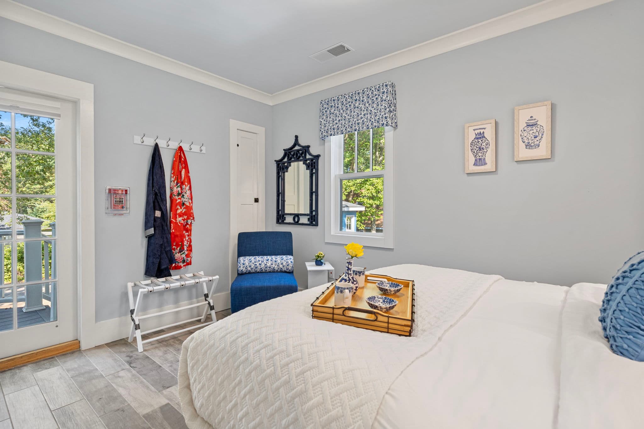 A cozy bedroom with light blue walls and wood-plank flooring featuring a bed with white linens and a wooden breakfast tray. The room includes a blue accent chair, a decorative black-framed mirror next to a window with a blue-and-white valance, and two framed blue-and-white prints on the wall. A glass door on the left leads to an outdoor balcony.