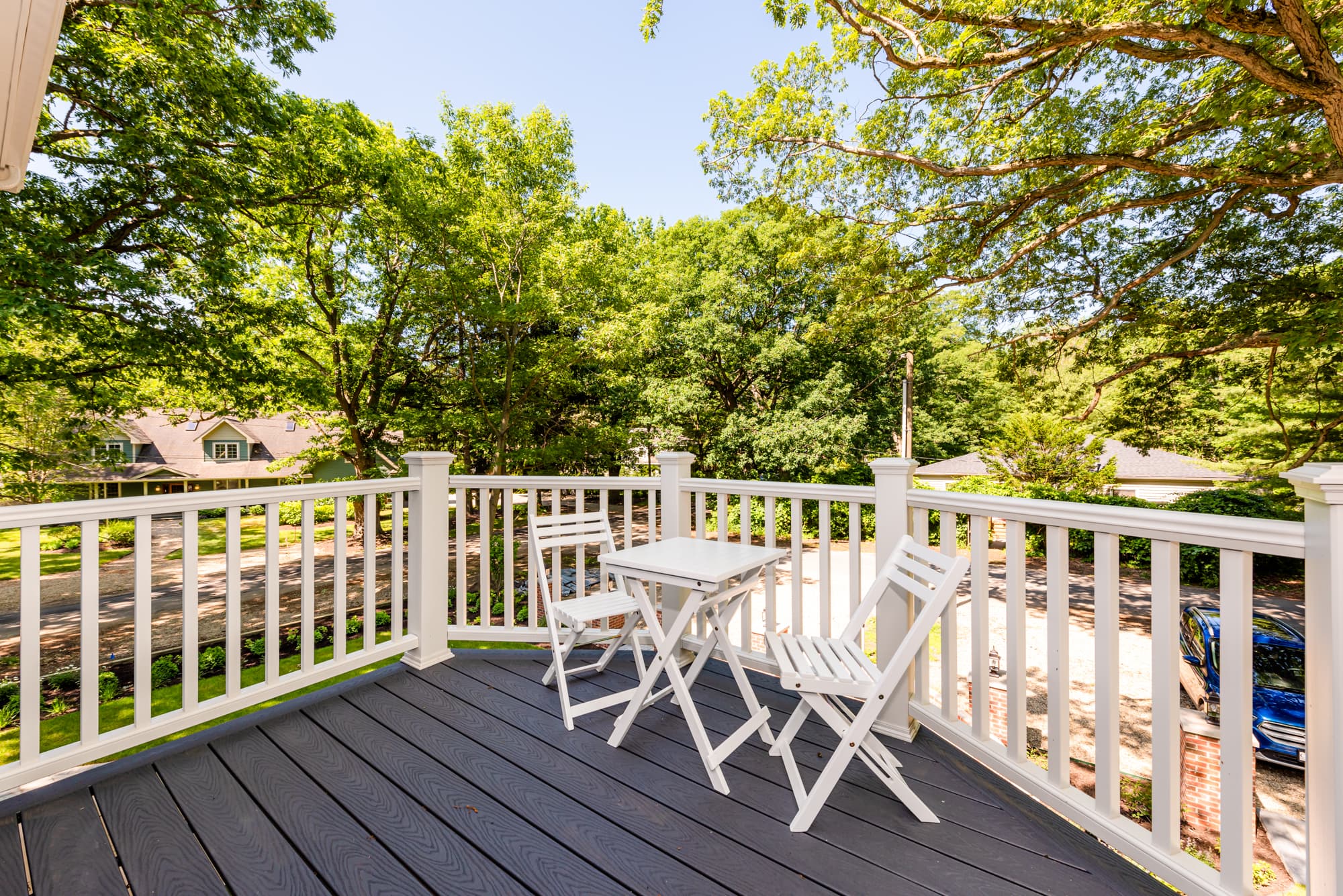 A spacious wooden deck with dark grey floorboards and white railings, featuring a small white folding table and two matching chairs. The deck is surrounded by lush green trees, providing a peaceful outdoor seating area.