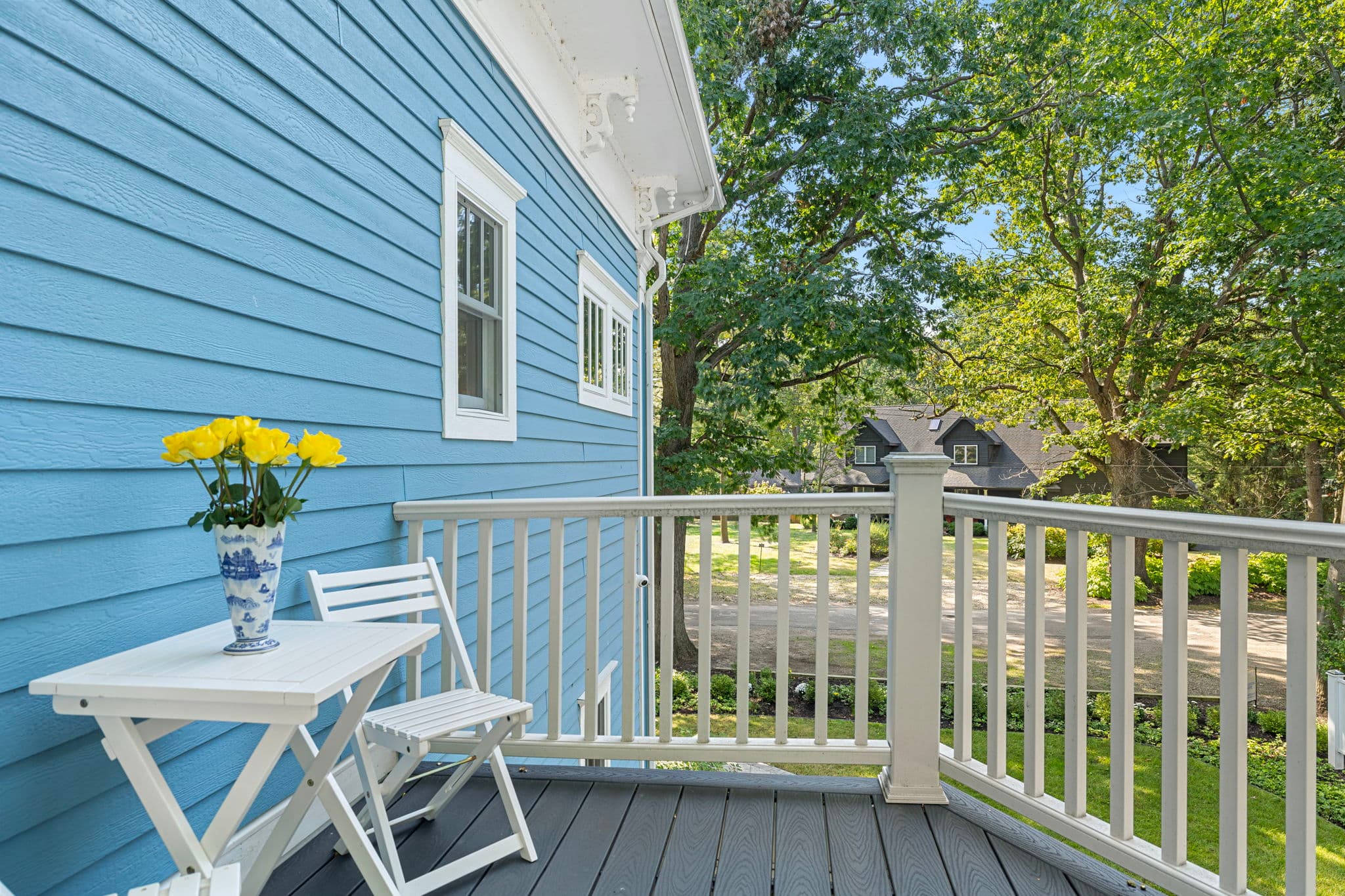 A close-up view of an outdoor deck featuring light blue siding on the house and white railings. A white folding table with a blue-and-white vase of yellow flowers and a matching white folding chair are positioned on the grey floorboards, with a view of a tree-lined street in the background.