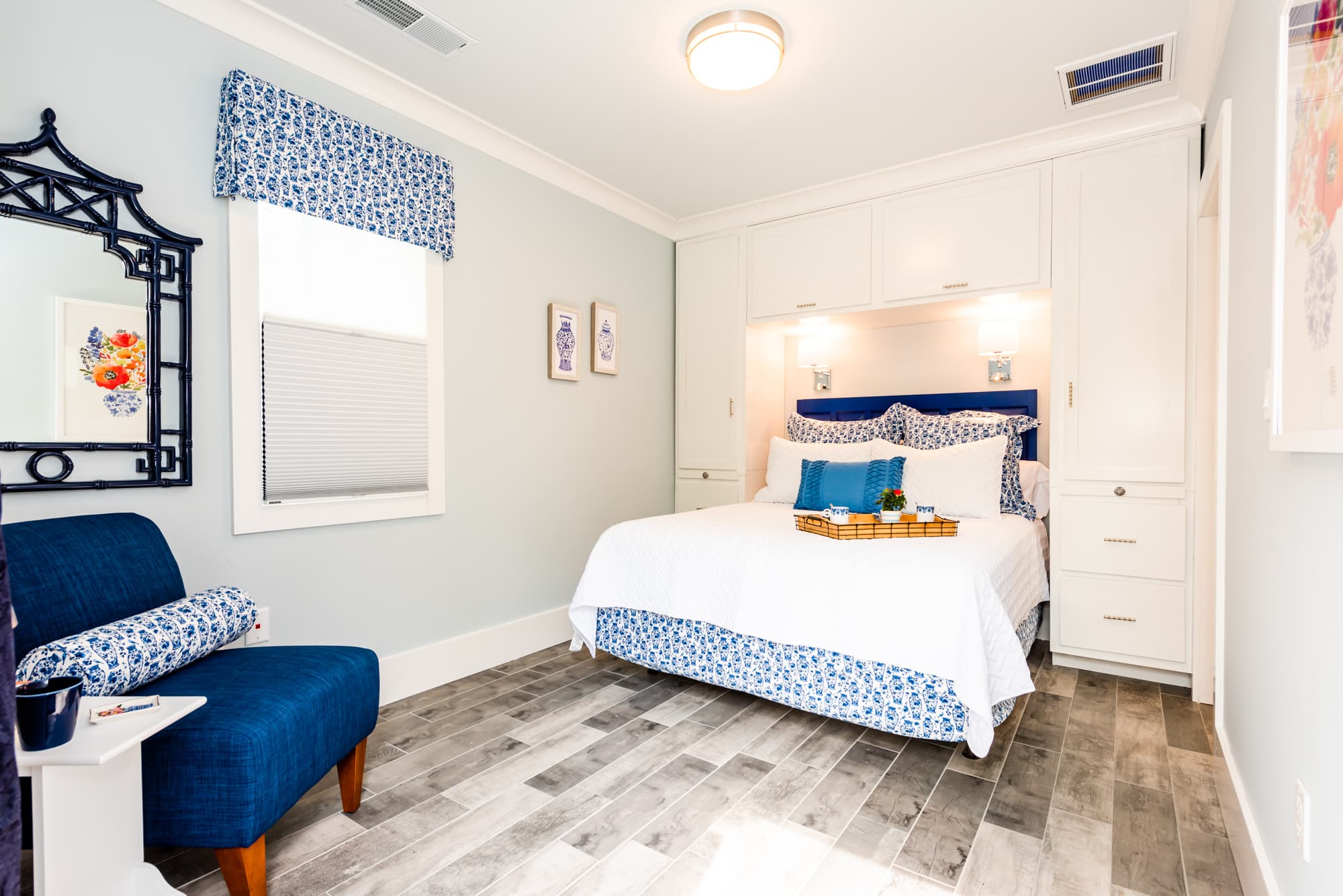 A cozy bedroom with light blue walls and wood-plank flooring features a bed tucked into a white built-in storage unit with cabinets and drawers on either side. The bed is dressed in white and blue patterned linens, complemented by a blue velvet headboard. To the left, a navy blue armchair with a patterned bolster pillow sits beneath a window with a blue-and-white patterned valance and an ornate black-framed mirror.