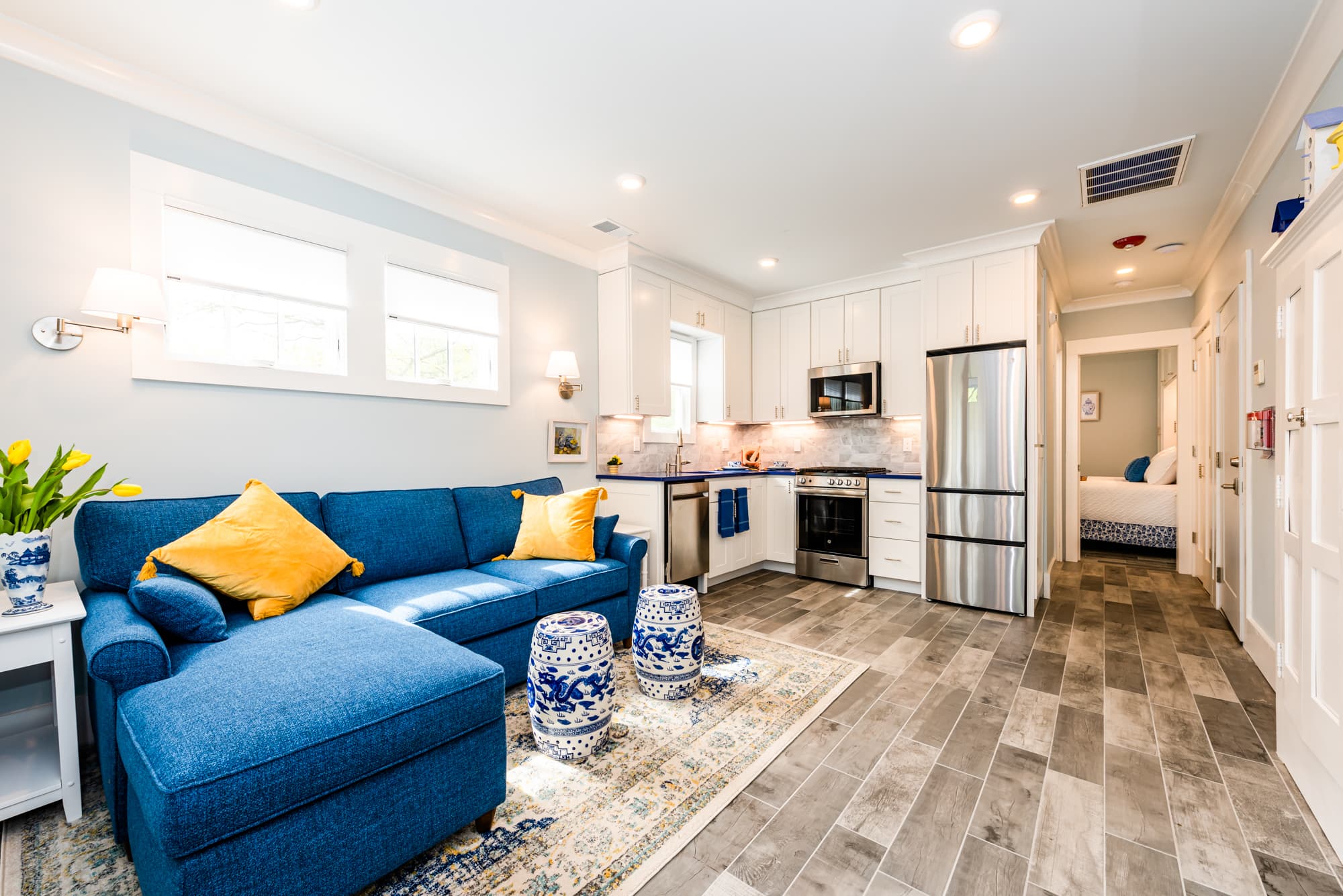 An open-concept living room and kitchen featuring a vibrant cobalt blue L-shaped sofa with yellow throw pillows. The kitchen area includes white cabinetry, stainless steel appliances, and a blue countertop. Two blue and white ceramic garden stools sit on a patterned rug in front of the sofa, and wood-plank flooring runs throughout the space.