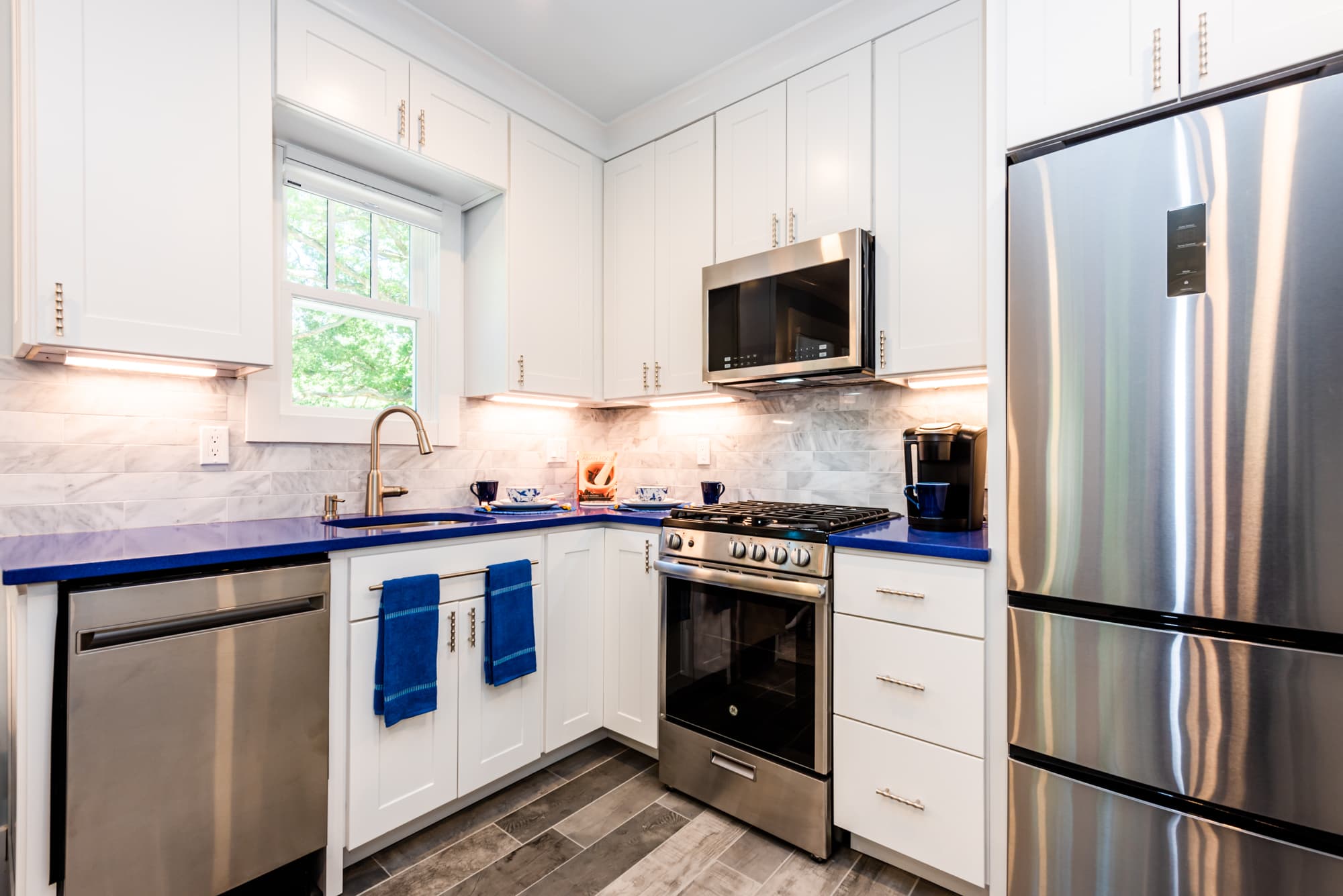 A modern kitchen with white cabinetry and a striking blue countertop. The space features stainless steel appliances, including a refrigerator, stove, microwave, and dishwasher, complemented by a marble-patterned backsplash and wood-look flooring.