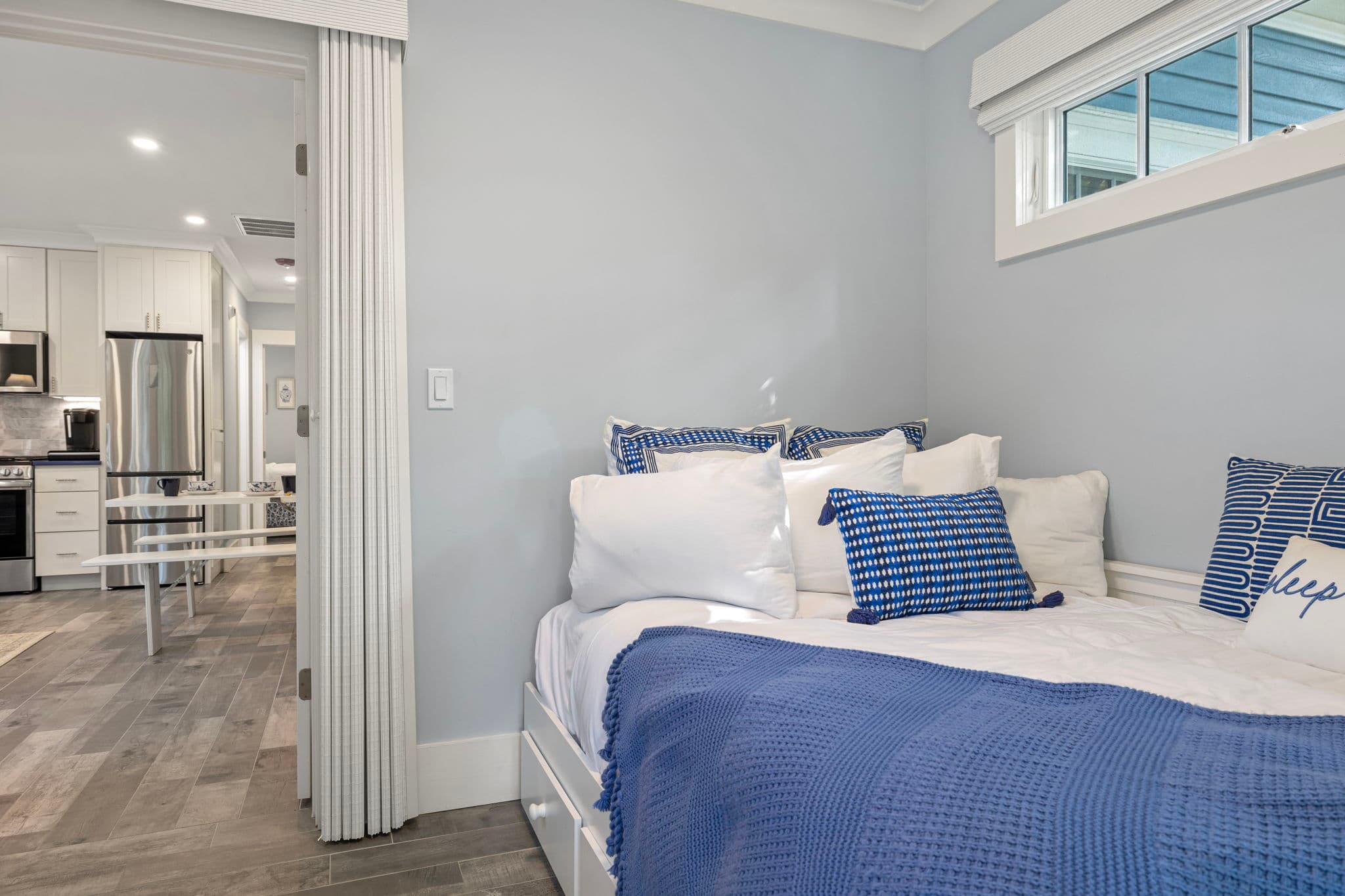 A small bedroom with light grey walls and wood-plank flooring featuring a white daybed. The bed is layered with white linens, blue-and-white patterned pillows, and a solid blue throw blanket. Through an open doorway, a modern kitchen with white cabinetry and stainless steel appliances is visible in the distance.