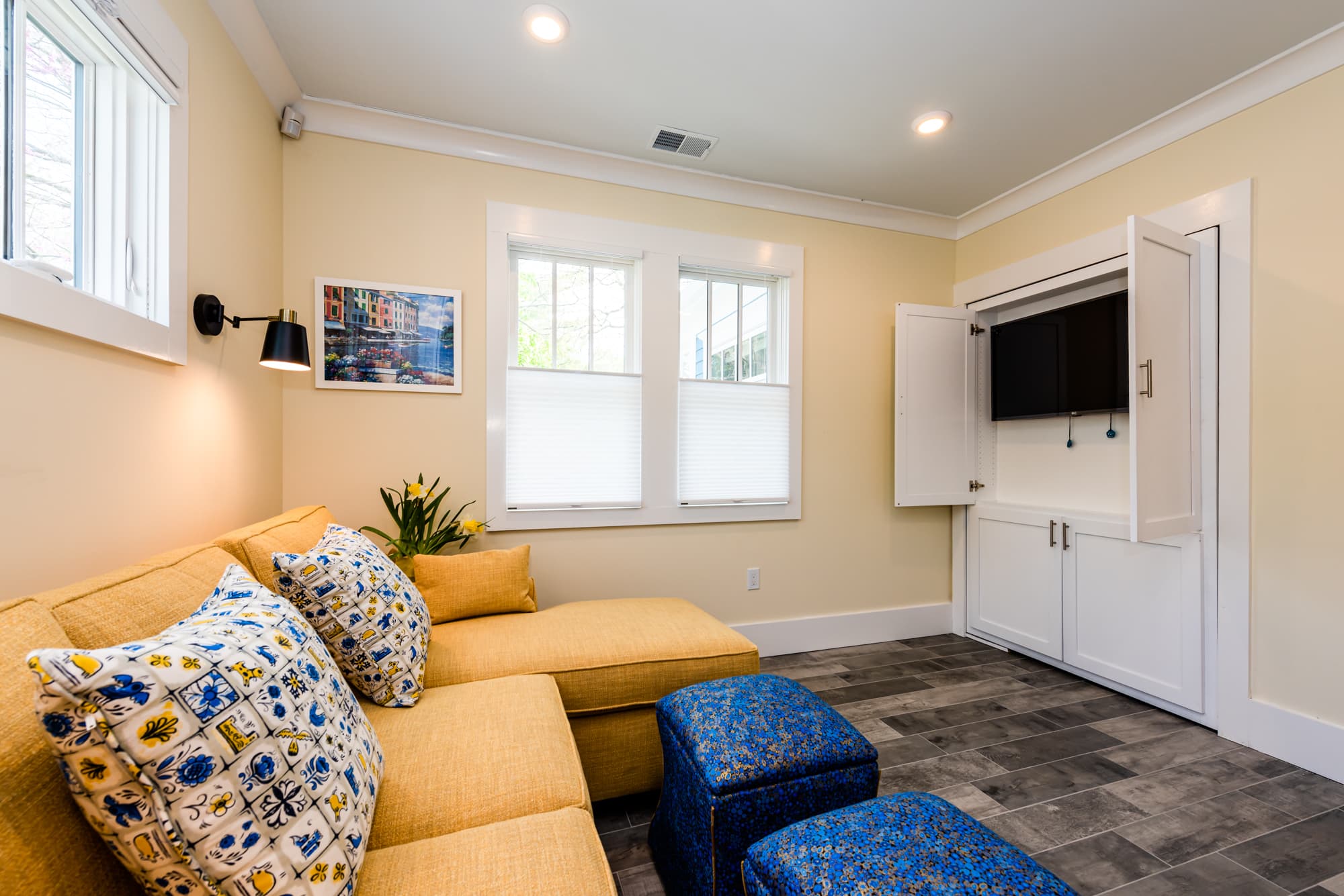 A cozy living area featuring a yellow sectional sofa with blue patterned pillows and two blue ottomans. Against the far wall, a white built-in cabinet with open doors reveals a flat-screen television, all set within a room with pale yellow walls and grey wood-plank flooring.