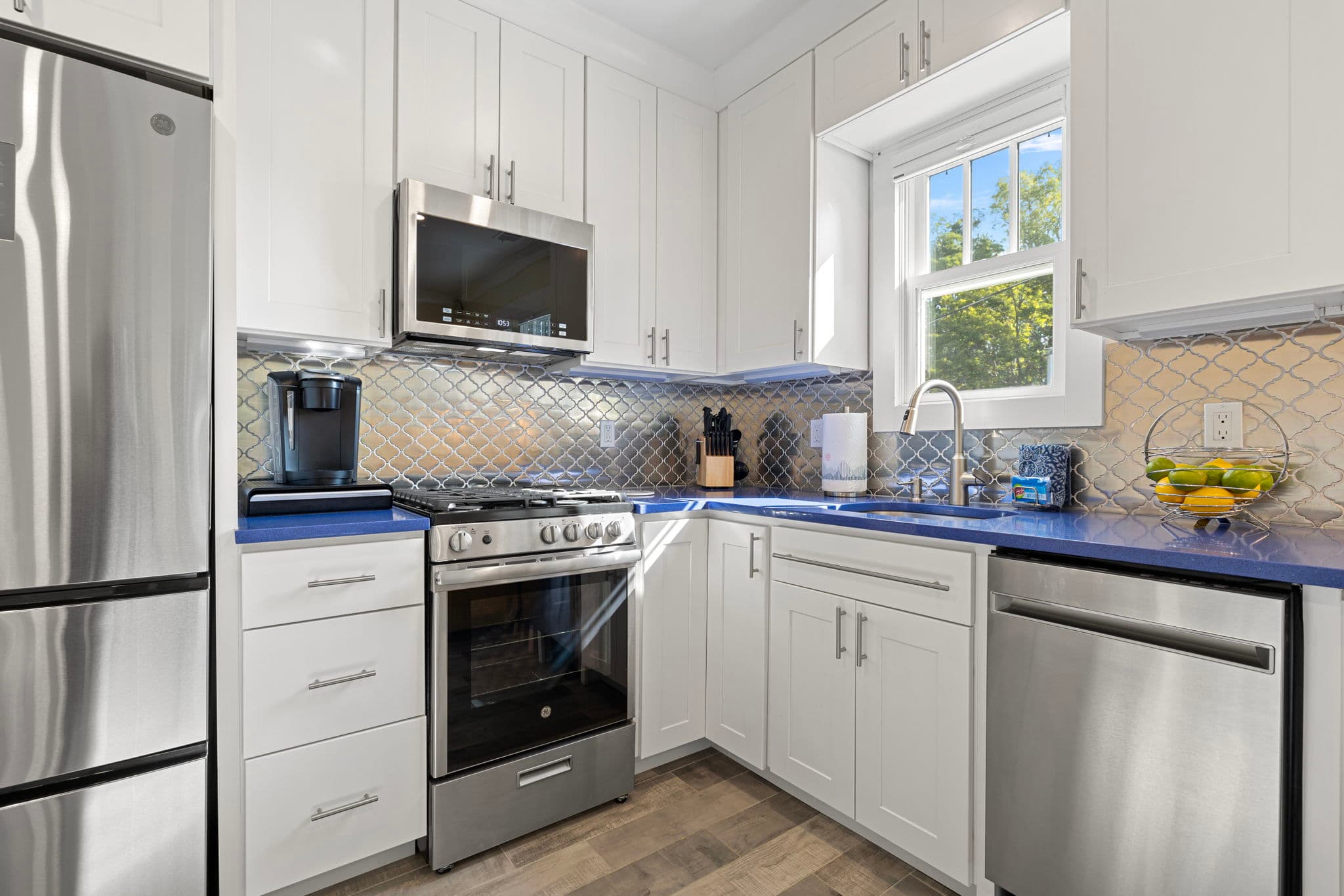 A close-up view of a modern kitchen featuring white shaker-style cabinetry, stainless steel appliances, and vibrant blue countertops. The space is finished with a neutral tile backsplash, a large window above the sink, and grey wood-plank flooring.