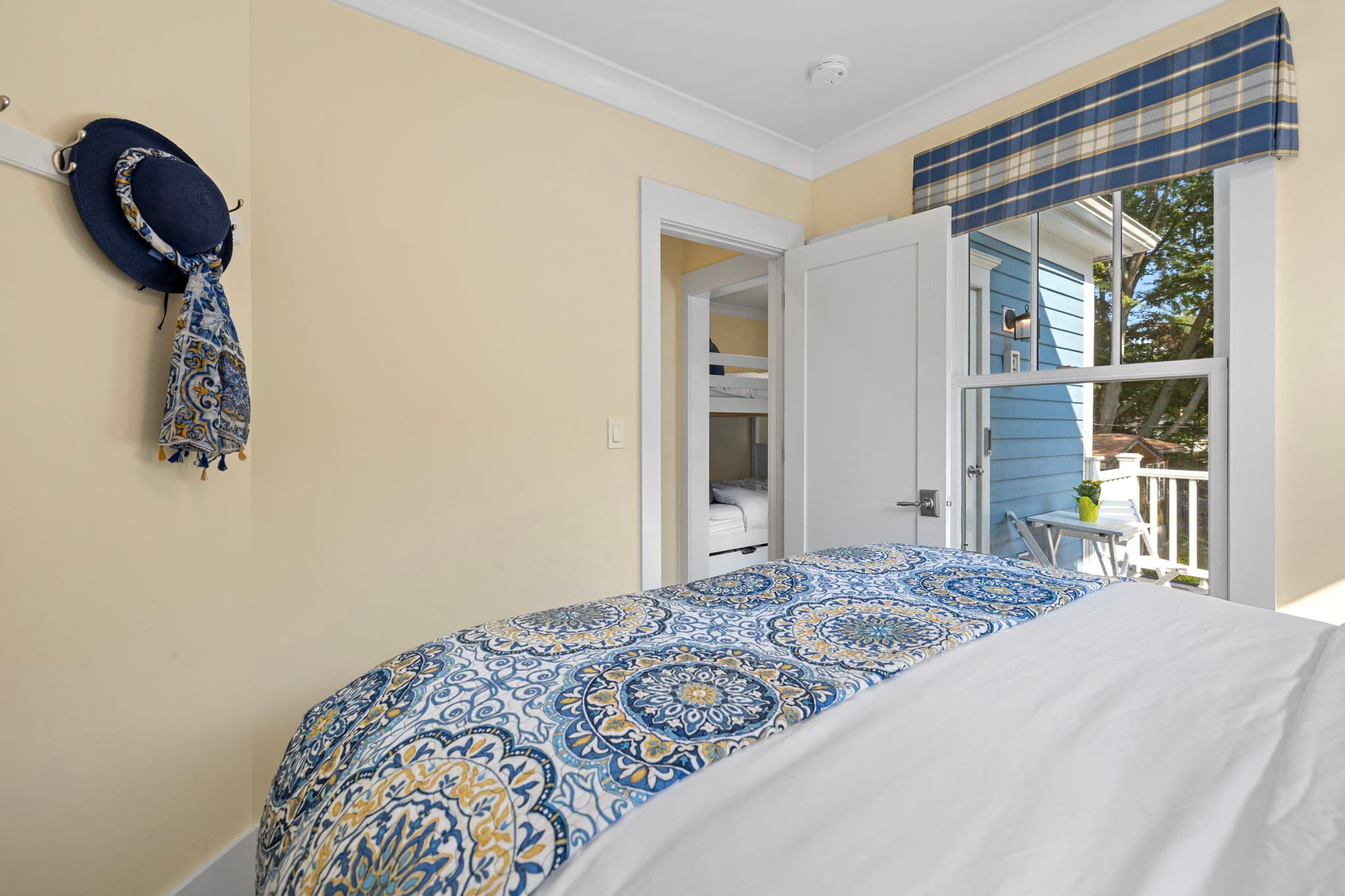 A bedroom view looking toward an open white door that reveals a glimpses of a bunk bed in the next room. The bed in the foreground is covered with a blue and yellow mandala-patterned runner, and a window to the right shows a view of a sunny blue-sided deck through a blue and white plaid valance.