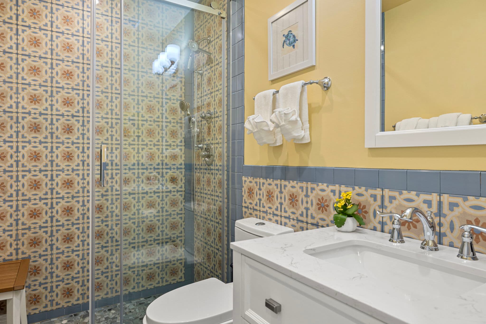 A vibrant bathroom featuring a glass-enclosed walk-in shower with ornate blue and orange patterned tiling on the back wall and floor. The space includes a white vanity with a marble top, a rectangular mirror, and a matching patterned tile backsplash that wraps around the yellow walls.