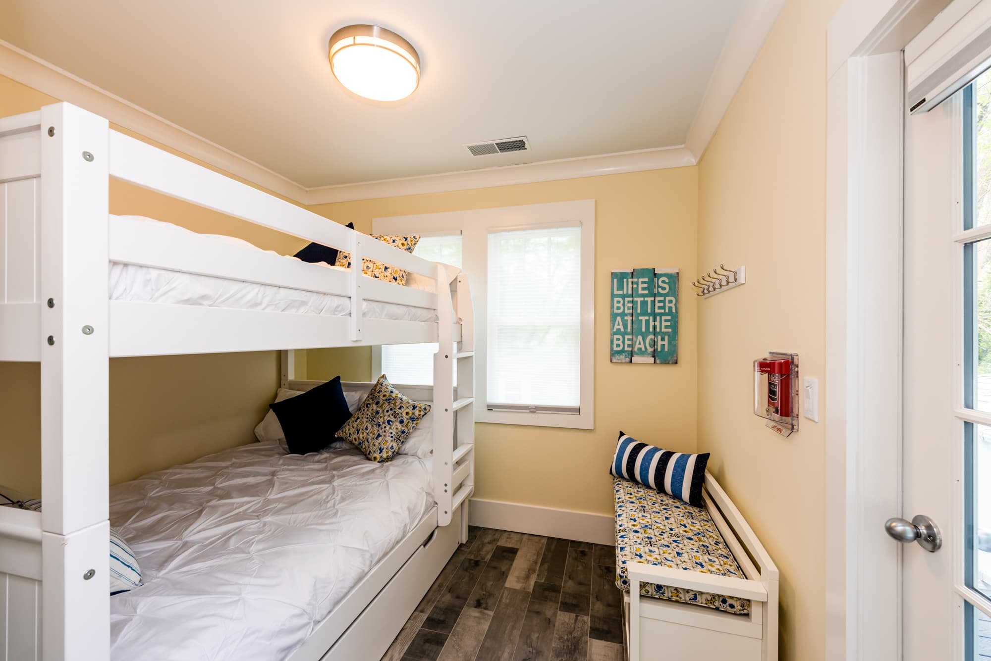 A small, cozy bedroom featuring white wooden bunk beds with navy and patterned throw pillows. The room has light yellow walls, grey wood-plank flooring, a small window with white trim, and a decorative "Life is Better at the Beach" sign on the wall next to a matching storage bench.