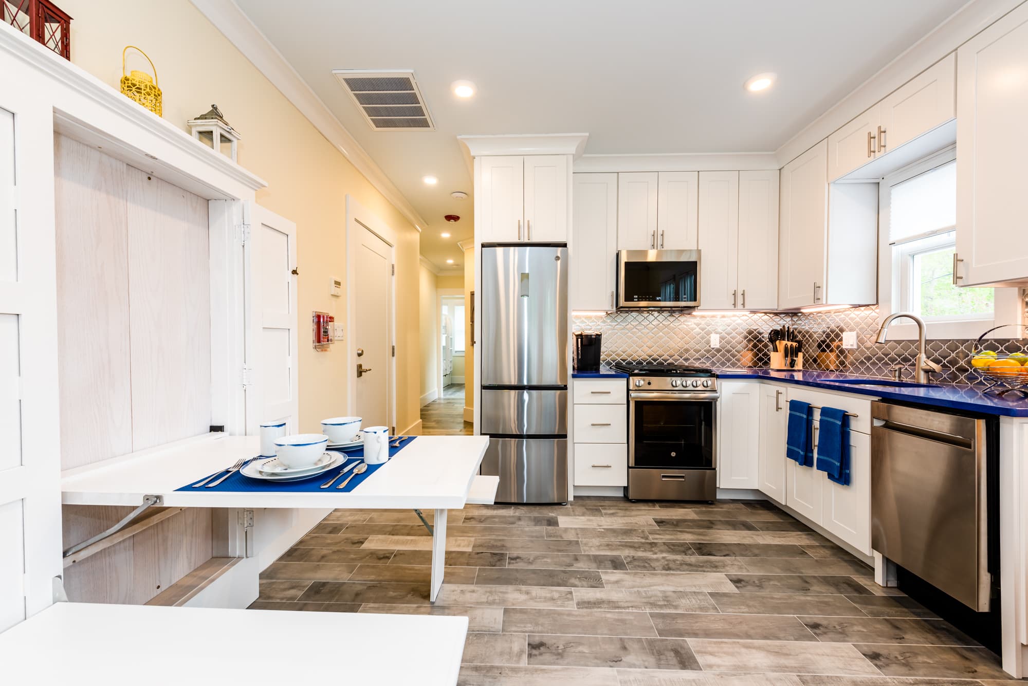 A modern kitchen and dining area featuring a built-in folding white table set with blue placemats and bowls. The kitchen in the background includes white cabinetry, a vibrant blue countertop, and stainless steel appliances.