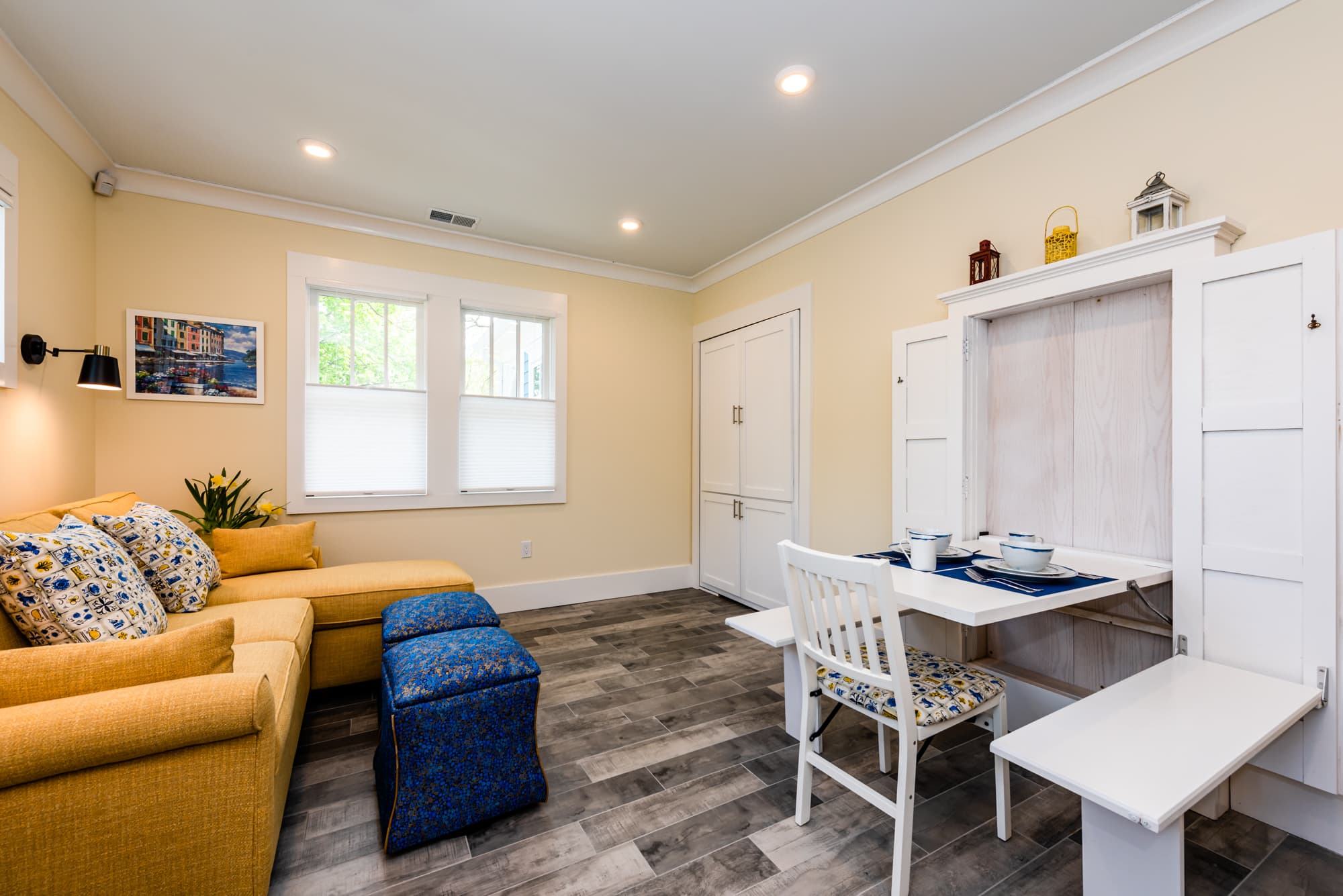 A versatile living and dining area featuring a yellow sectional sofa and blue patterned ottomans on the left. To the right, a white wall-mounted folding table is extended with place settings, accompanied by a white chair and a folding bench, all set against neutral walls and grey wood-plank flooring.