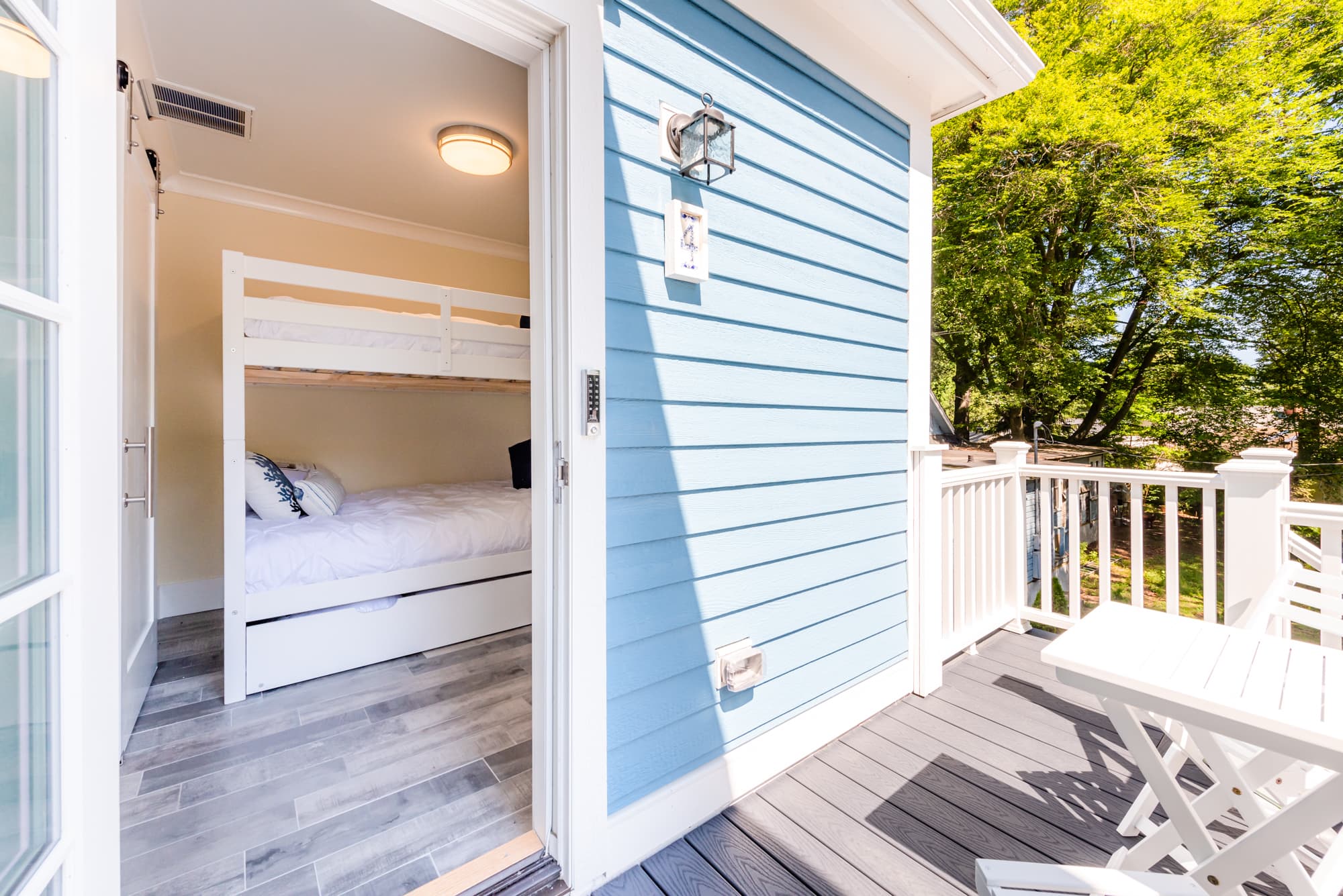 Exterior view from a grey wooden deck looking through an open door into a bright bedroom with white bunk beds. The house features light blue horizontal siding, and a white folding table is visible on the sunny deck surrounded by lush green trees.