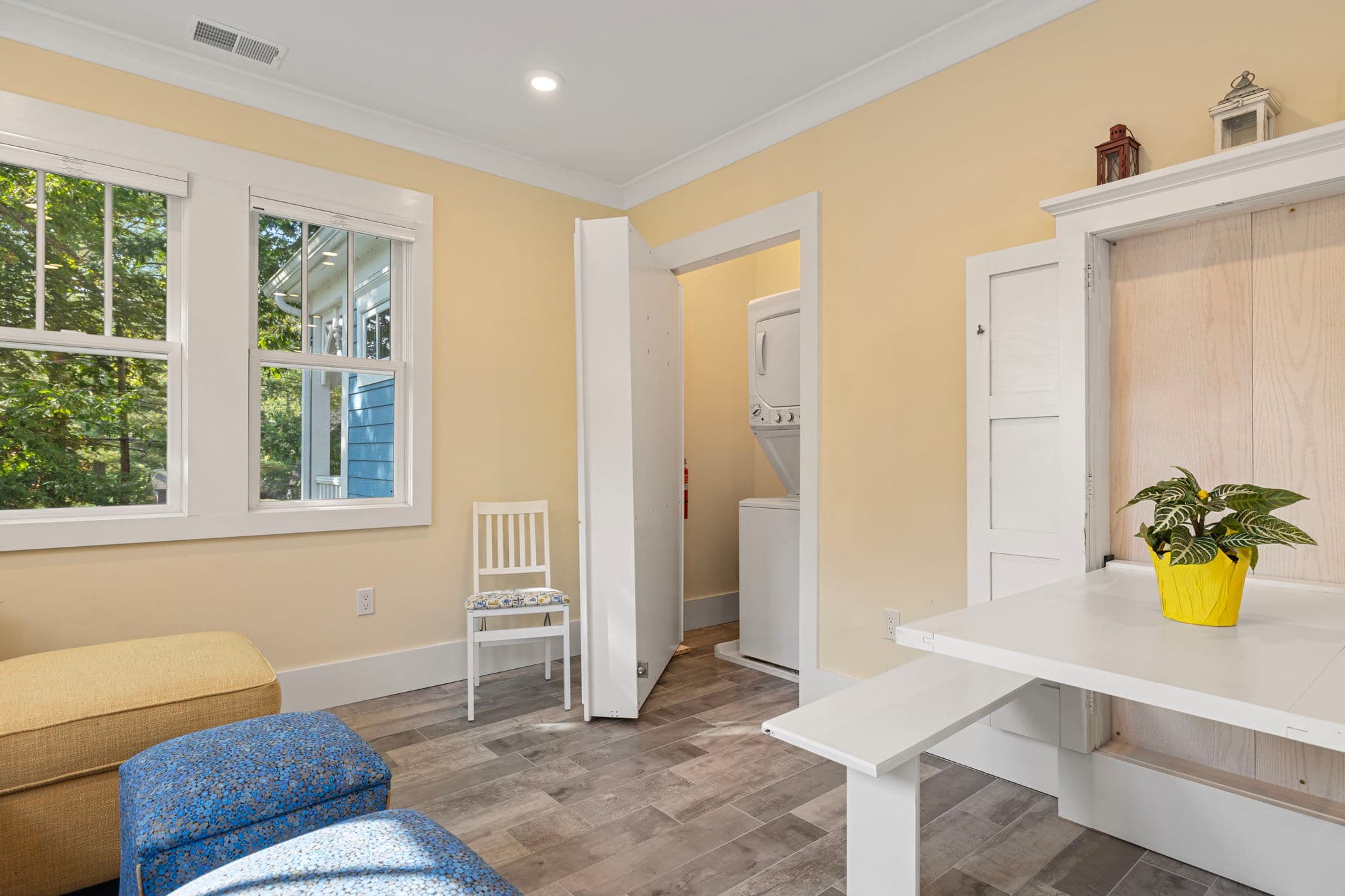 A sunlit multi-functional room featuring a white wall-mounted folding table with a small potted plant in a yellow bowl. In the background, a white bifold door is partially open, revealing a stacked washer and dryer unit in a laundry nook.