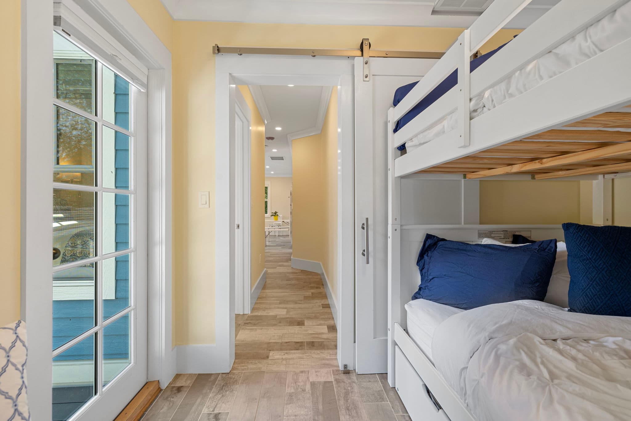 A view from inside the bunk bed room looking down a narrow hallway. To the right is the white wooden bunk bed with navy blue pillows, and to the left is a white-framed glass door leading to the exterior. A sliding white barn door stands open, revealing the continuation of the grey wood-plank flooring throughout the home.