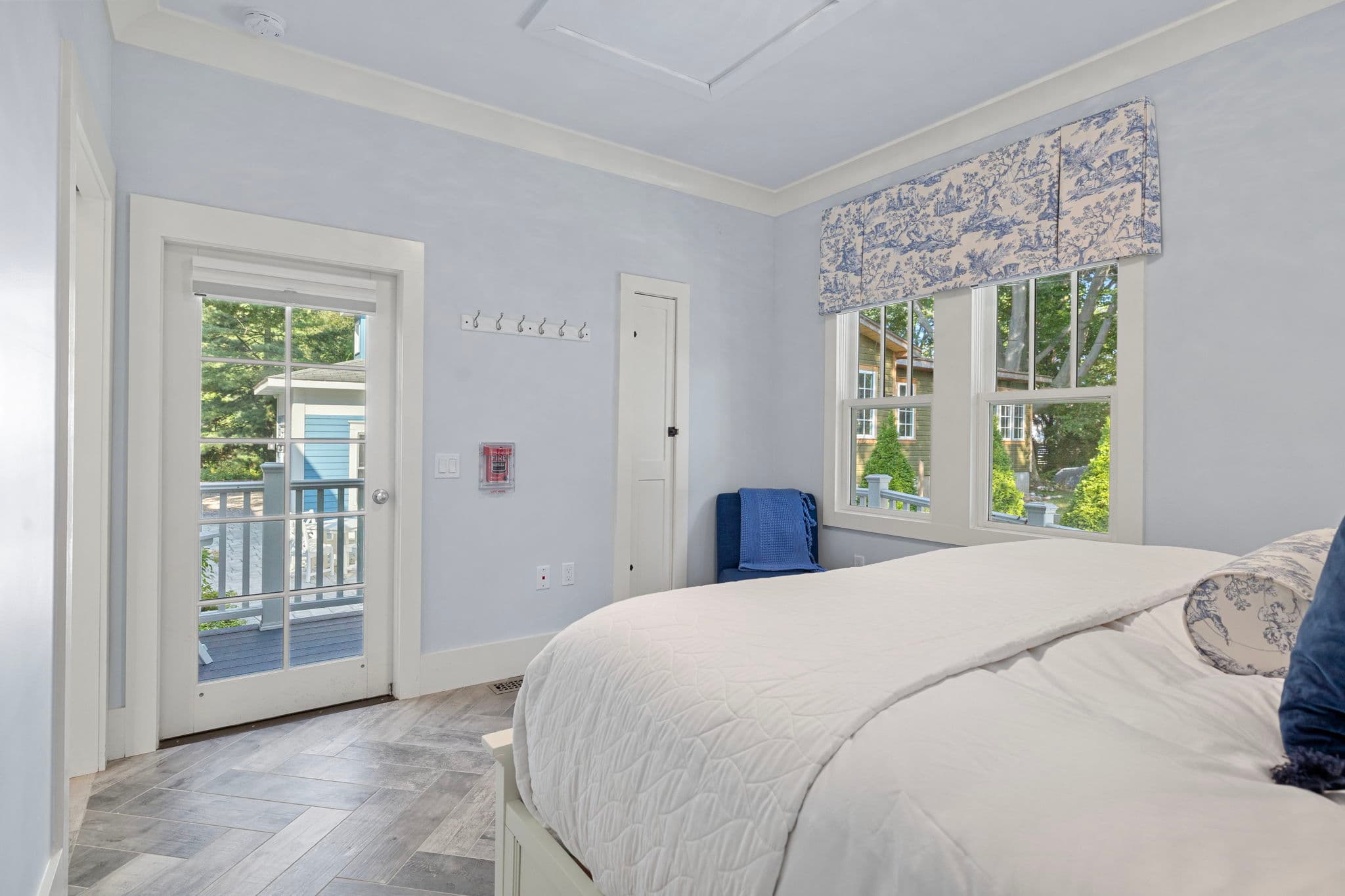 A bedroom featuring a white wooden bunk bed with a white comforter and navy blue throw pillows. The room has light yellow walls, grey wood-plank flooring, and a white-framed glass door that leads to an exterior deck. A small white storage bench with a blue and yellow patterned cushion sits against the wall next to a decorative "Life is Better at the Beach" sign.