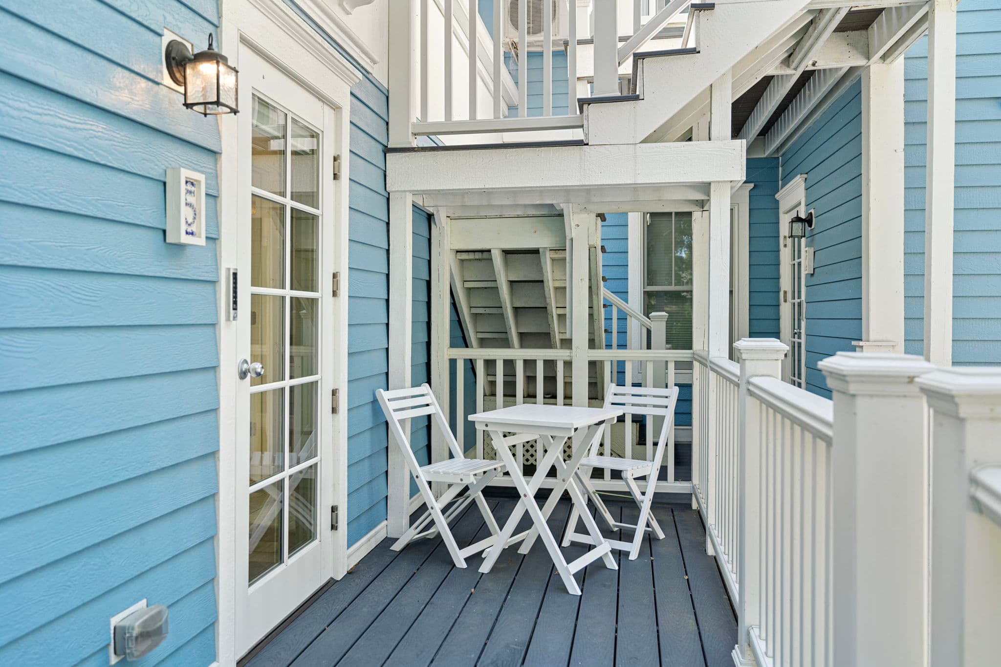 Exterior view of a small white wooden table and two folding chairs set on a dark grey deck. The area is enclosed by white railings and the light blue horizontal siding of the house, with a white-framed glass door providing access to the interior.