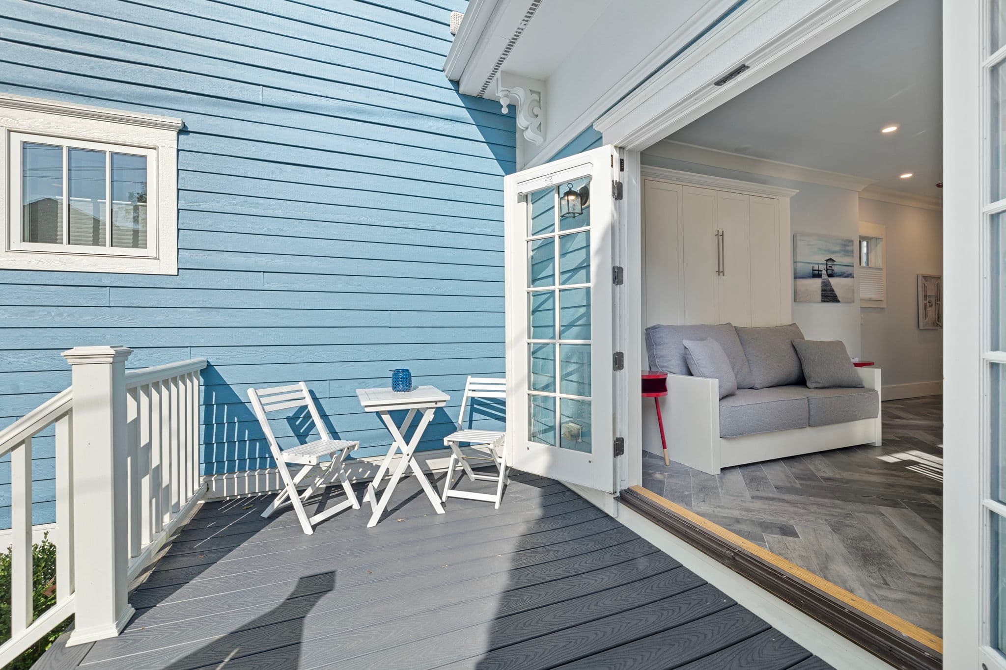 An exterior view from a grey wooden deck looking through wide-open French doors into a modern living area with a grey sofa and herringbone floors. The house features light blue siding, and a small white bistro set with two chairs is positioned on the sunny deck.
