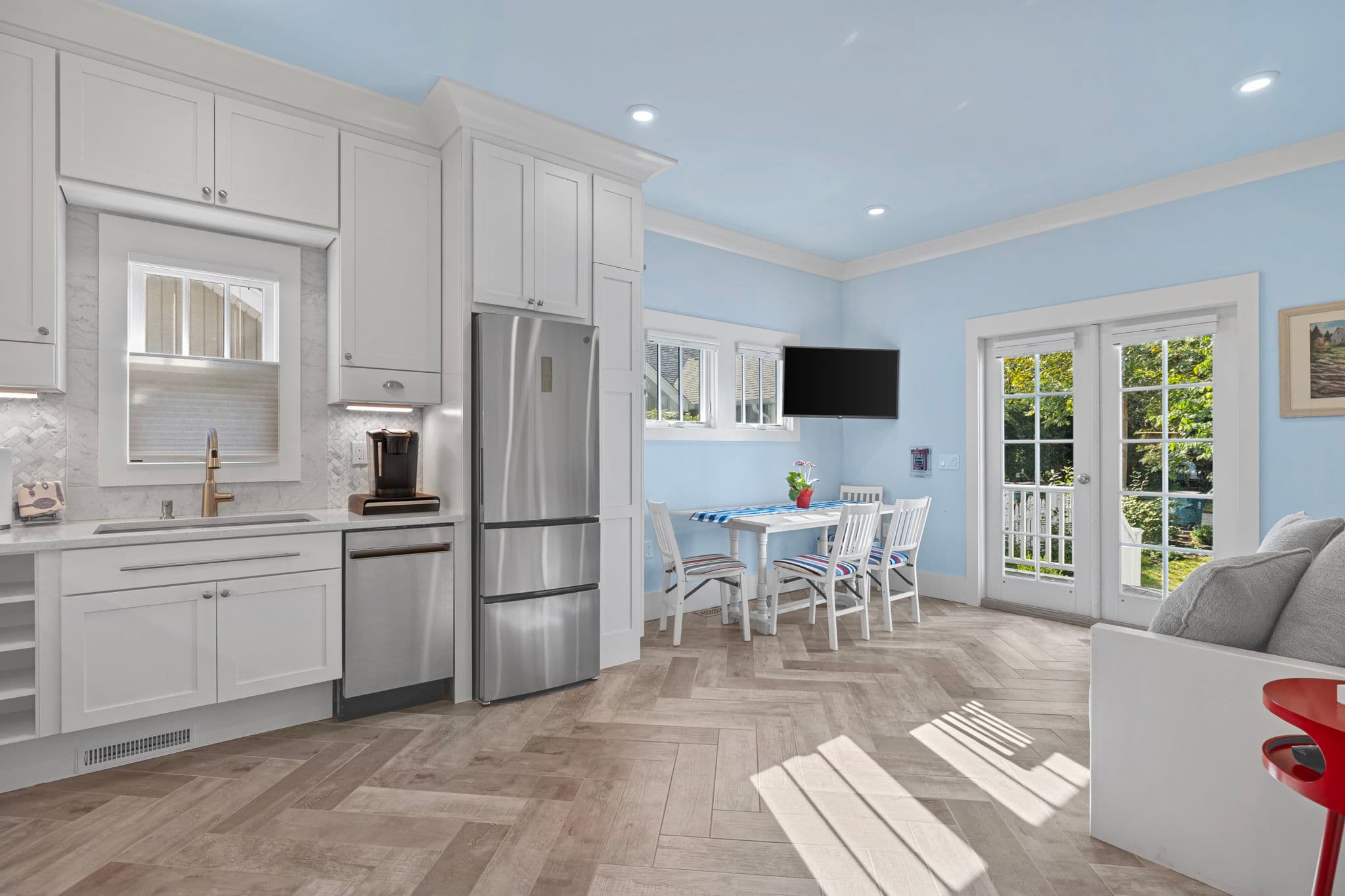 A bright, modern kitchen and dining area featuring white shaker-style cabinetry and a stainless steel refrigerator. To the right, a white dining table with striped chair cushions is set against a light blue wall with a wall-mounted TV, all atop grey herringbone flooring.