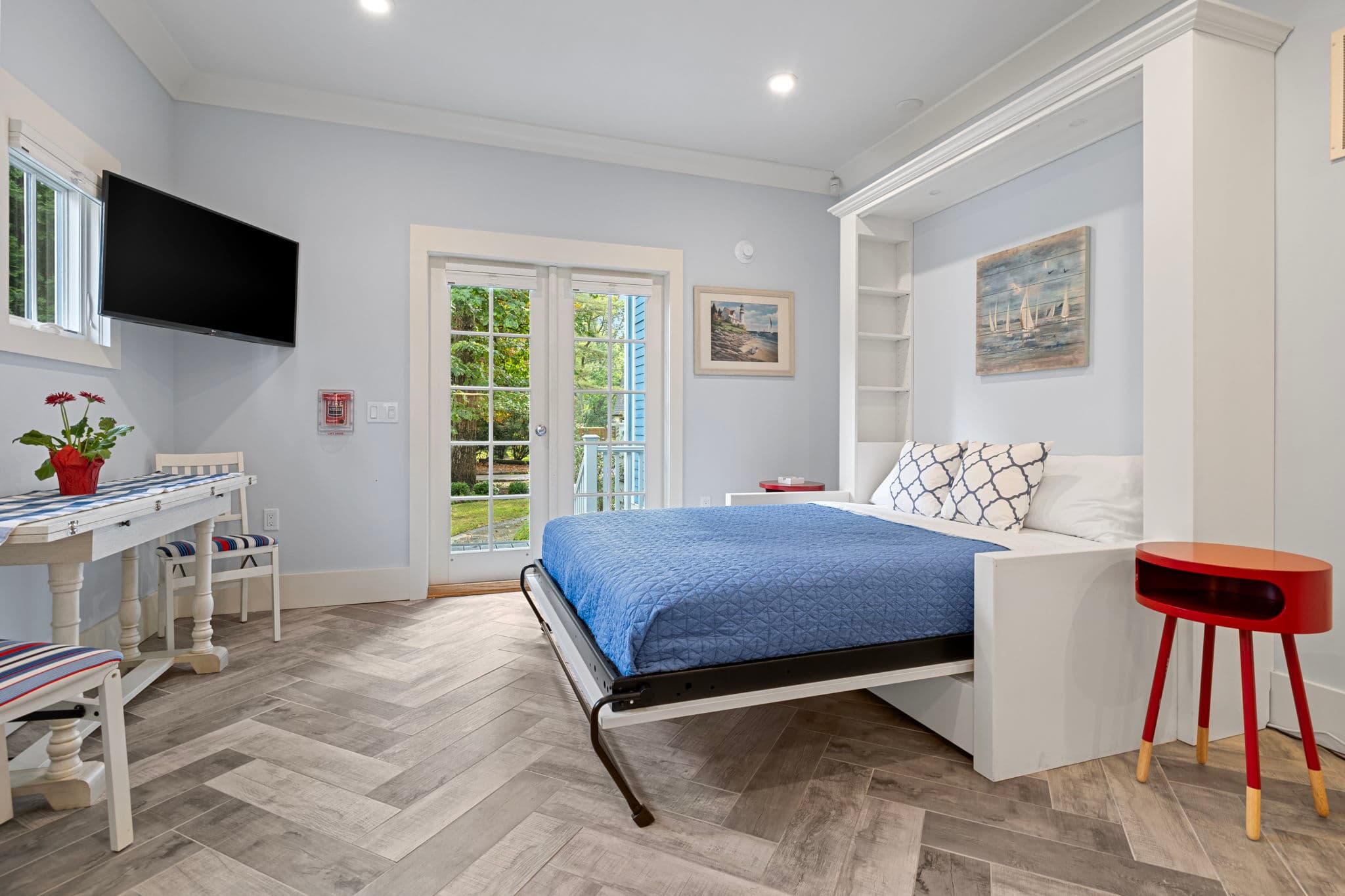 A versatile living area featuring a white Murphy bed with a blue comforter. The room includes grey herringbone-patterned flooring, light blue walls, a wall-mounted TV, and white French doors that open to a lush green outdoor space.