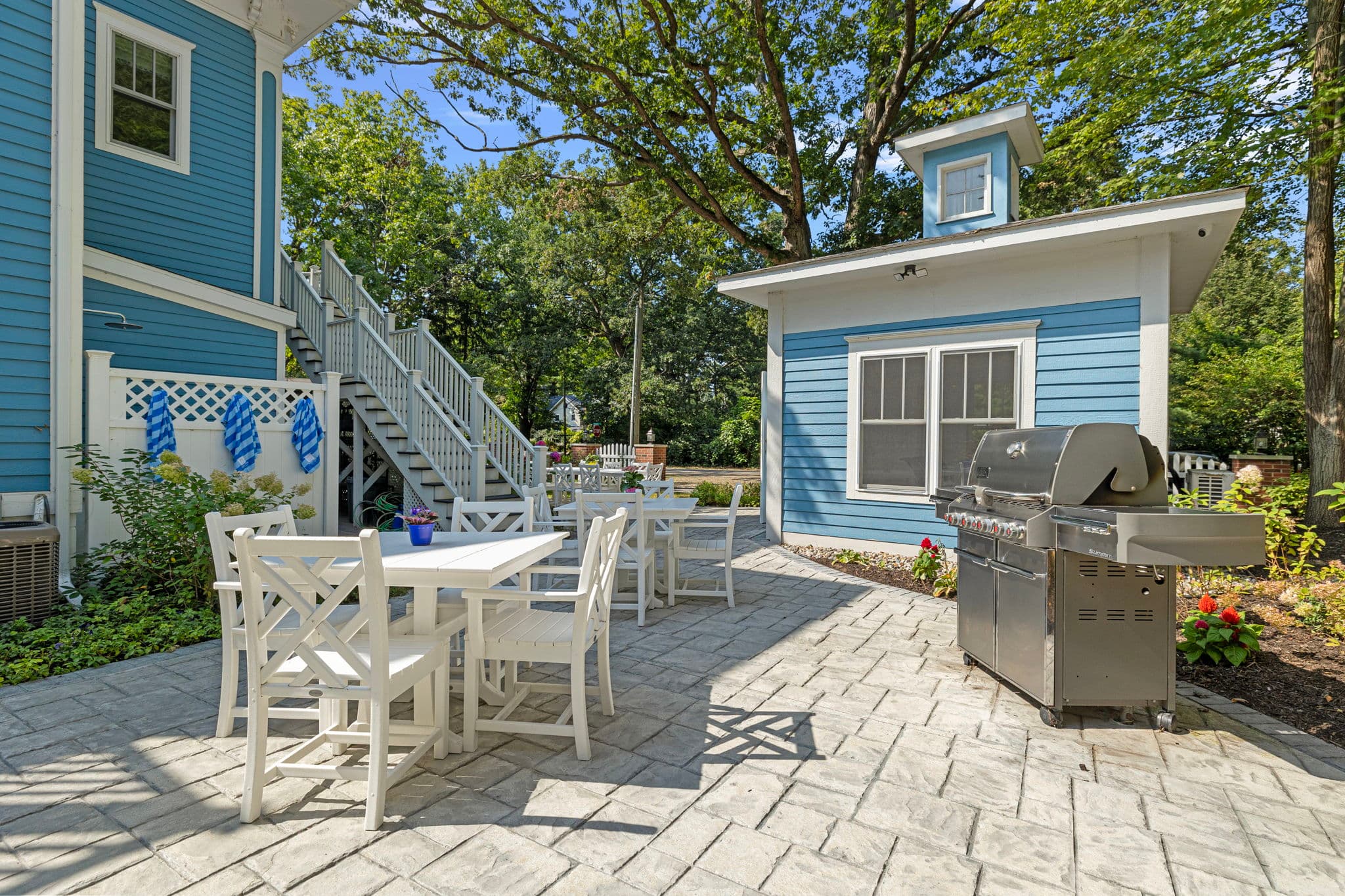 A spacious stone-paved patio area featuring multiple white outdoor dining sets and a stainless steel gas grill in the foreground. The yard includes a blue outbuilding with a small cupola and a portion of the main vibrant blue house with an exterior staircase, all surrounded by mature green trees.