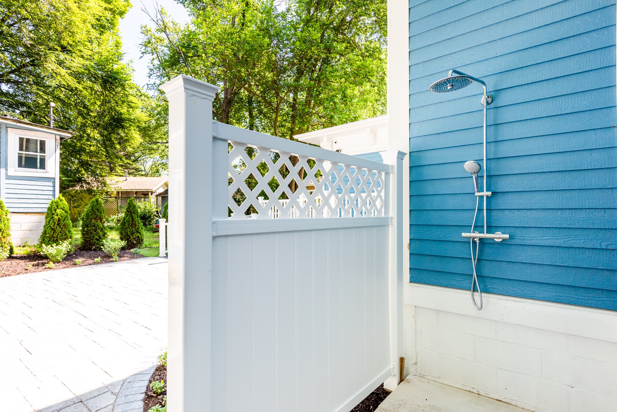 An outdoor shower area featuring a stainless steel showerhead and handheld attachment mounted against vibrant blue horizontal siding. The space is partially enclosed by a white privacy fence with a lattice-top detail, overlooking a sunny paved patio and lush green trees in the background.