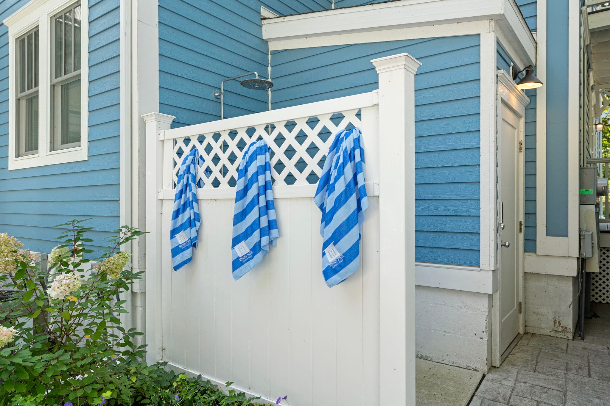 A white privacy fence with a lattice top features three blue and white striped towels hanging from it. The fence is positioned against the vibrant blue horizontal siding of the house, next to a white door with an overhead light and a glimpse of the outdoor shower area.