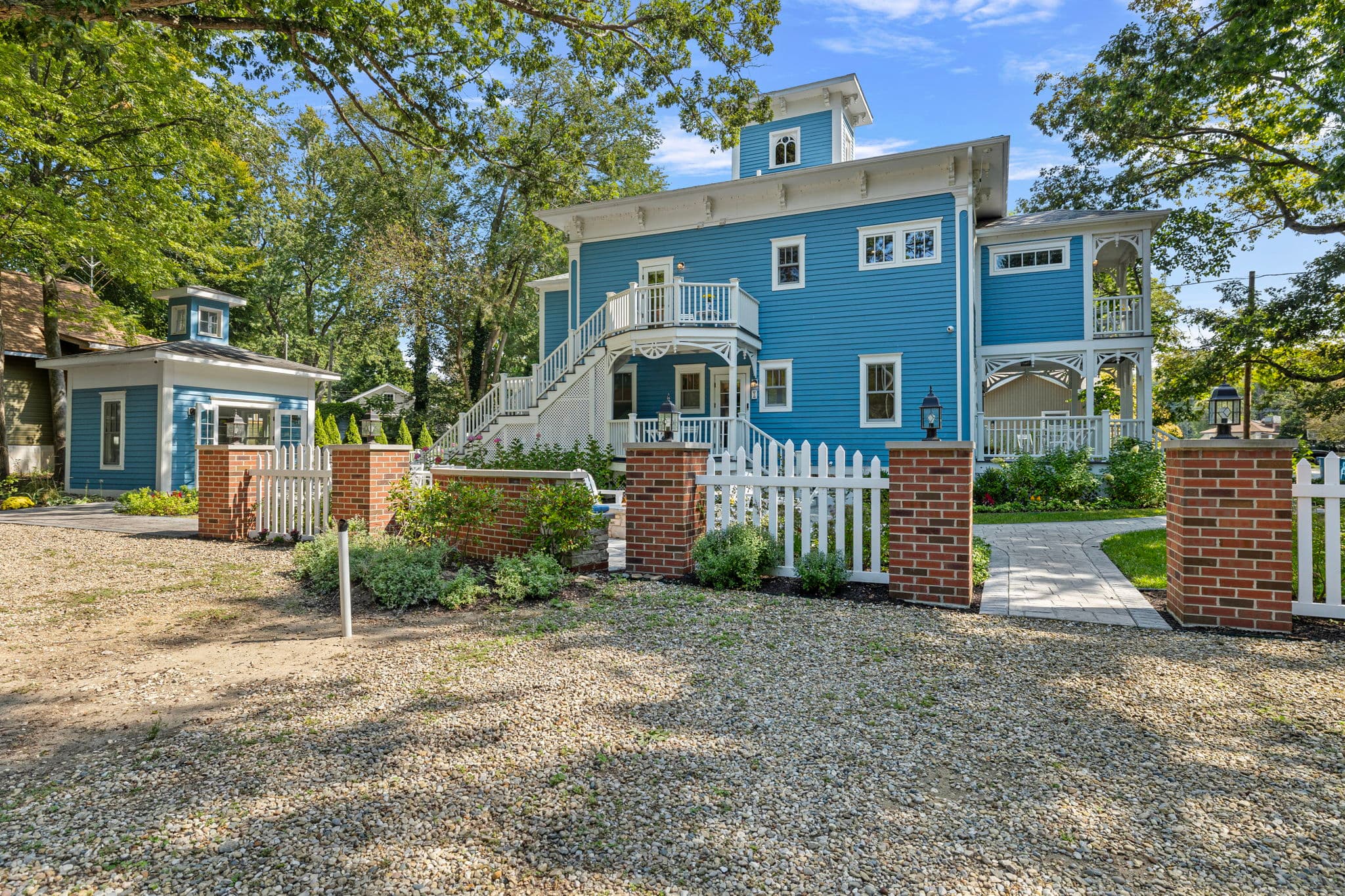 An exterior view of the Union Pier Summer House property, featuring the three-story blue house and matching garden pavilion from behind a white picket fence. The house has extensive white-railed porches and a staircase leading to the yard, while brick pillars frame the gated entrance to the stone-paved walkways and lush lawn.