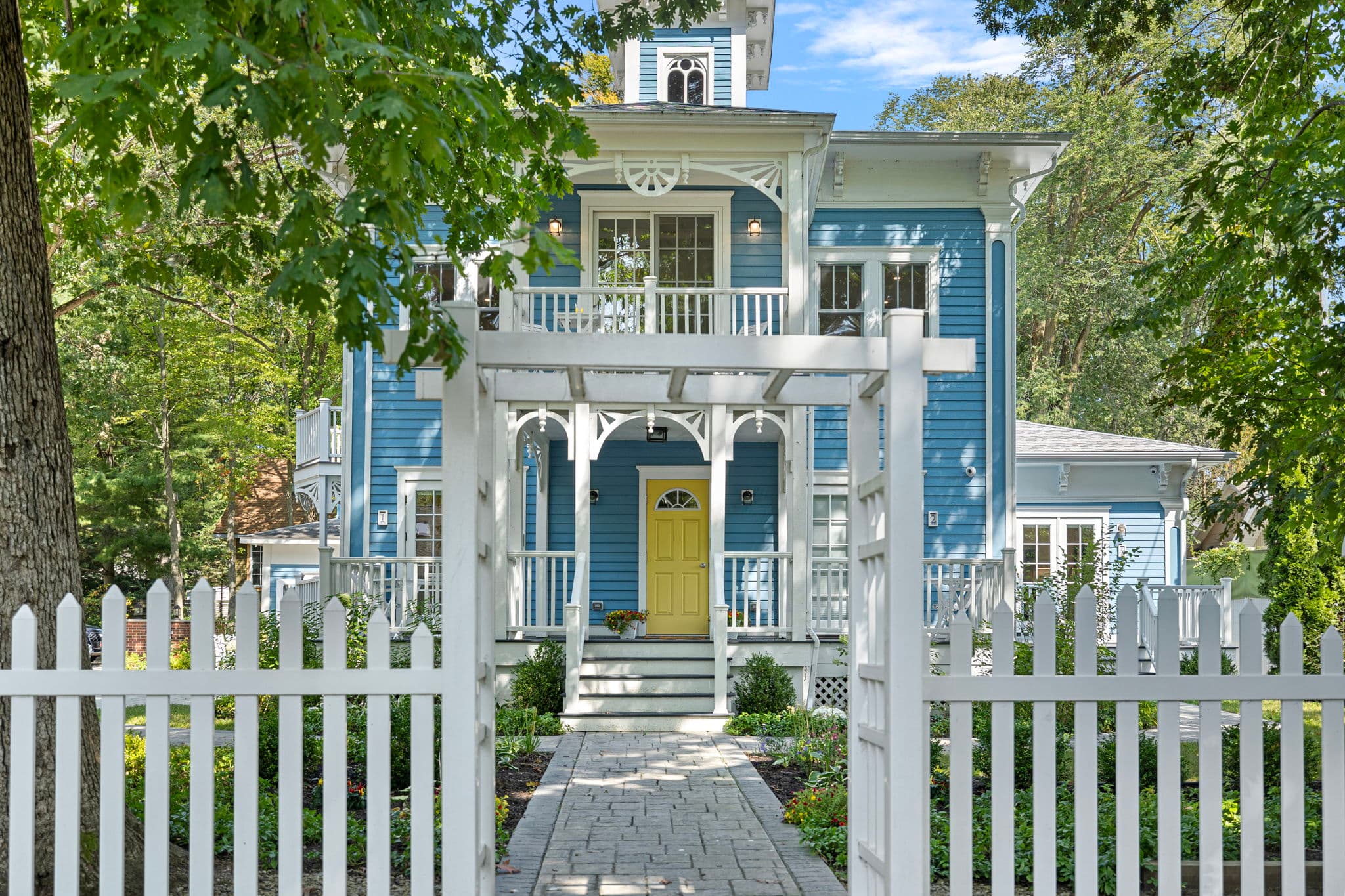 A view from the front yard looking through a white wooden pergola and picket fence toward the Union Pier Summer House. The house features vibrant blue siding, ornate white trim on its two levels of porches, and a bright yellow front door centered at the top of a short set of stairs.