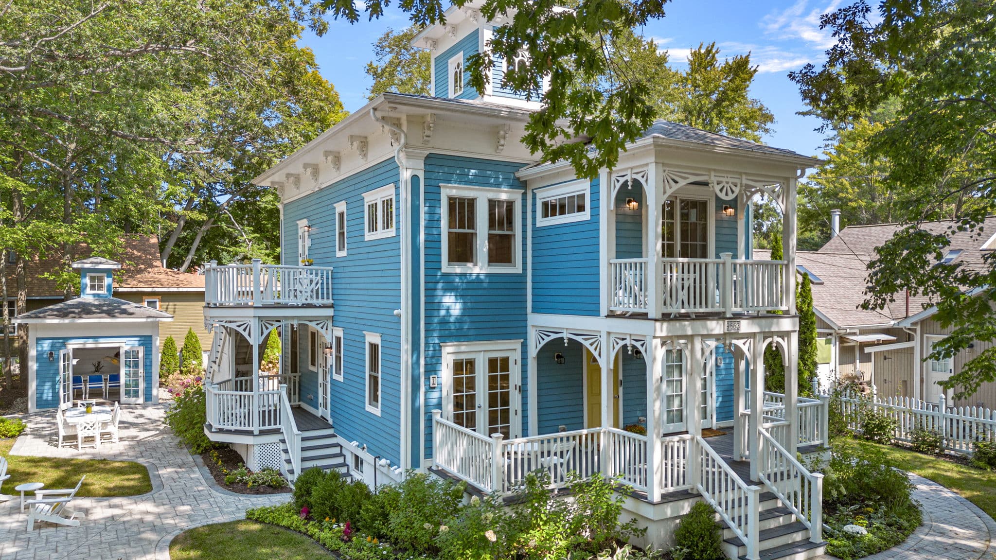 A three-story house with vibrant blue siding and white ornate trim, featuring multiple wrap-around porches and balconies with white railings. The home is surrounded by lush green trees and a landscaped yard with stone pathways and a matching blue outbuilding.