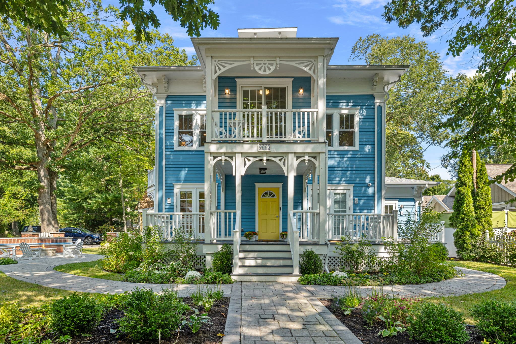A symmetrical front view of the Union Pier Summer House, featuring vibrant blue horizontal siding, ornate white trim, and a bright yellow front door. The house has two levels of white-railed porches with decorative columns, surrounded by a lush green lawn and stone-paved pathways.