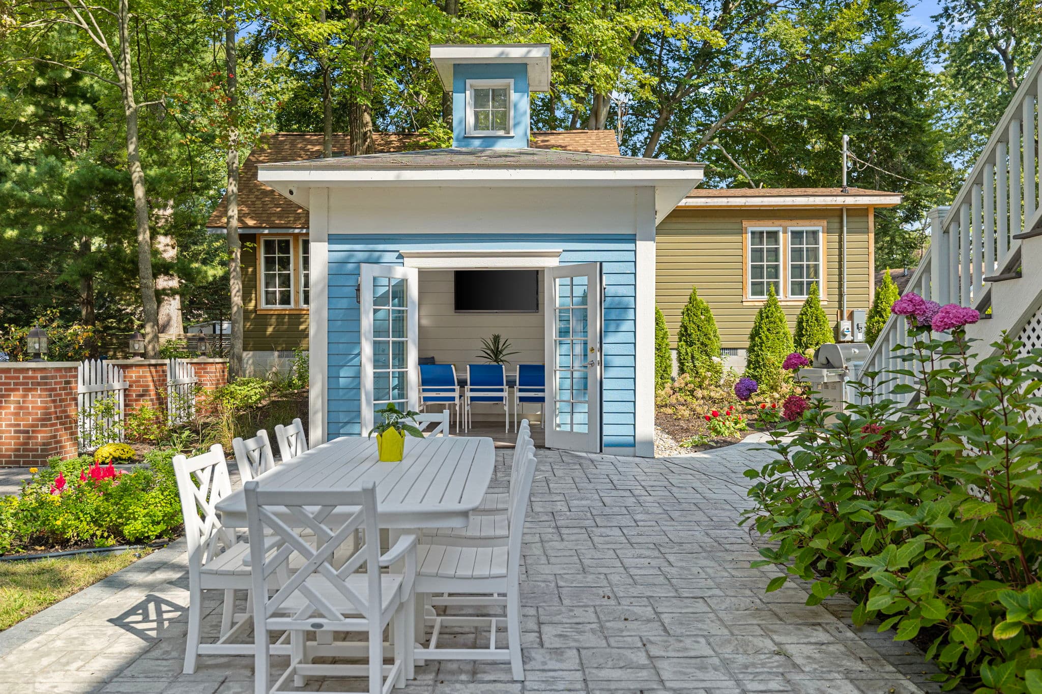 A landscaped stone patio area featuring a white outdoor dining table and chairs in the foreground. In the background, a small blue outbuilding with open French doors reveals an interior with a wall-mounted TV, while lush green trees and garden beds surround the space.